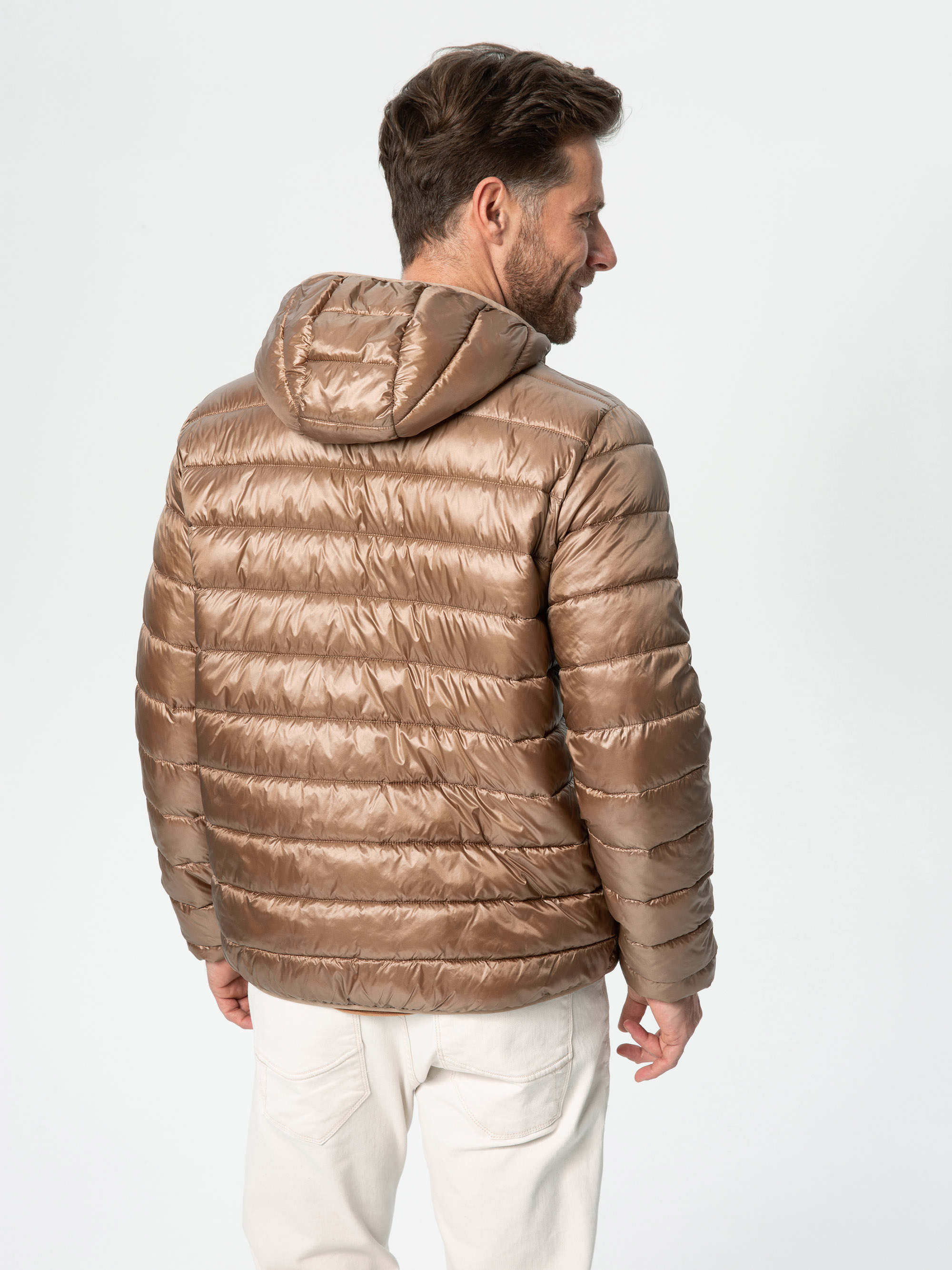 Ein stehender Mann, der eine glänzende, hellbraune Pufferjacke mit Kapuze trägt, gepaart mit einer cremefarbenen Hose, vor einem schlichten hellen Hintergrund.