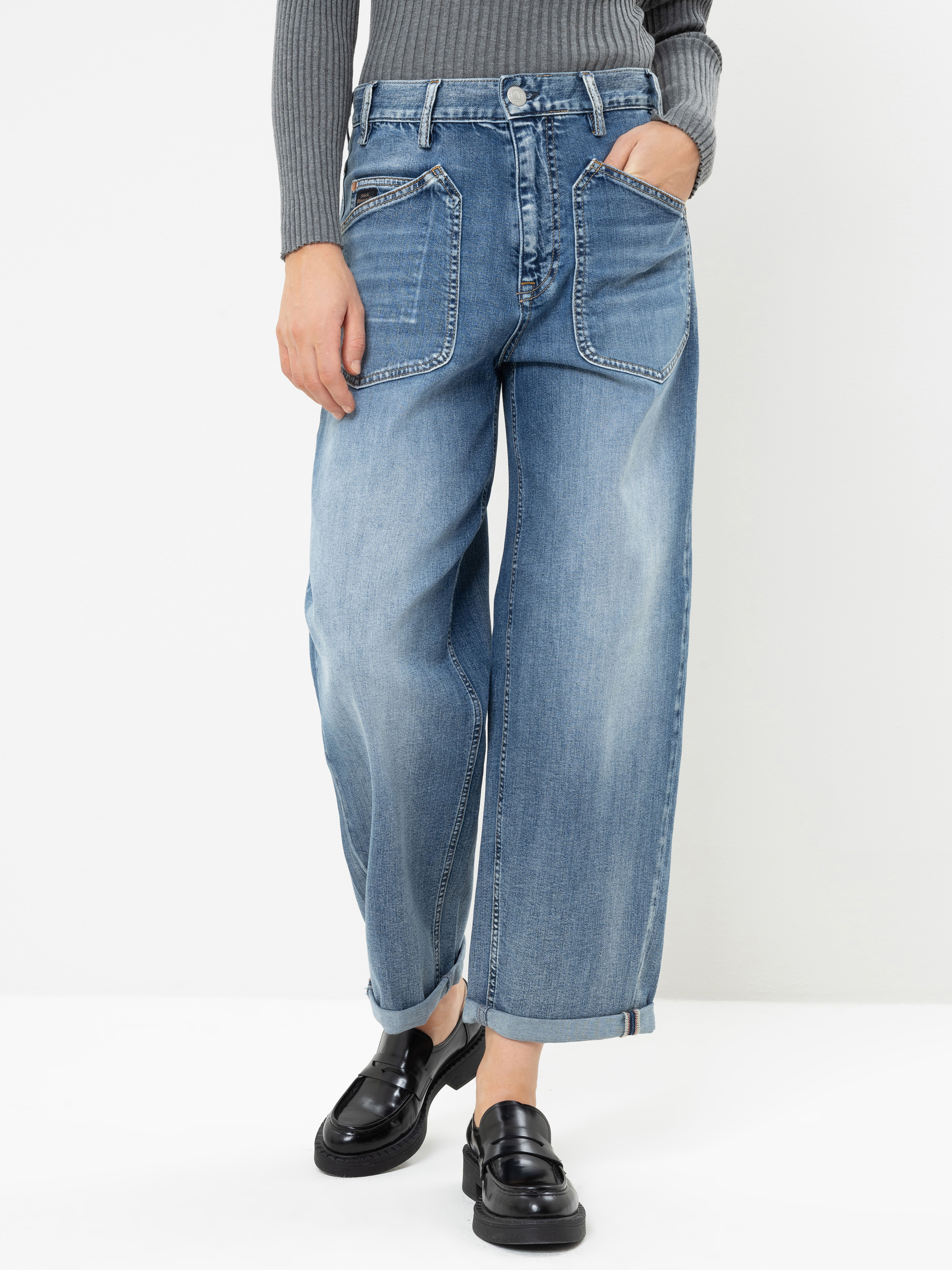Eine Person, die hellblaue Jeans mit weitem Bein und großen Vordertaschen, ein graues, geripptes Langarm-Oberteil und schwarze Loafer trägt, steht vor einem weißen Hintergrund.