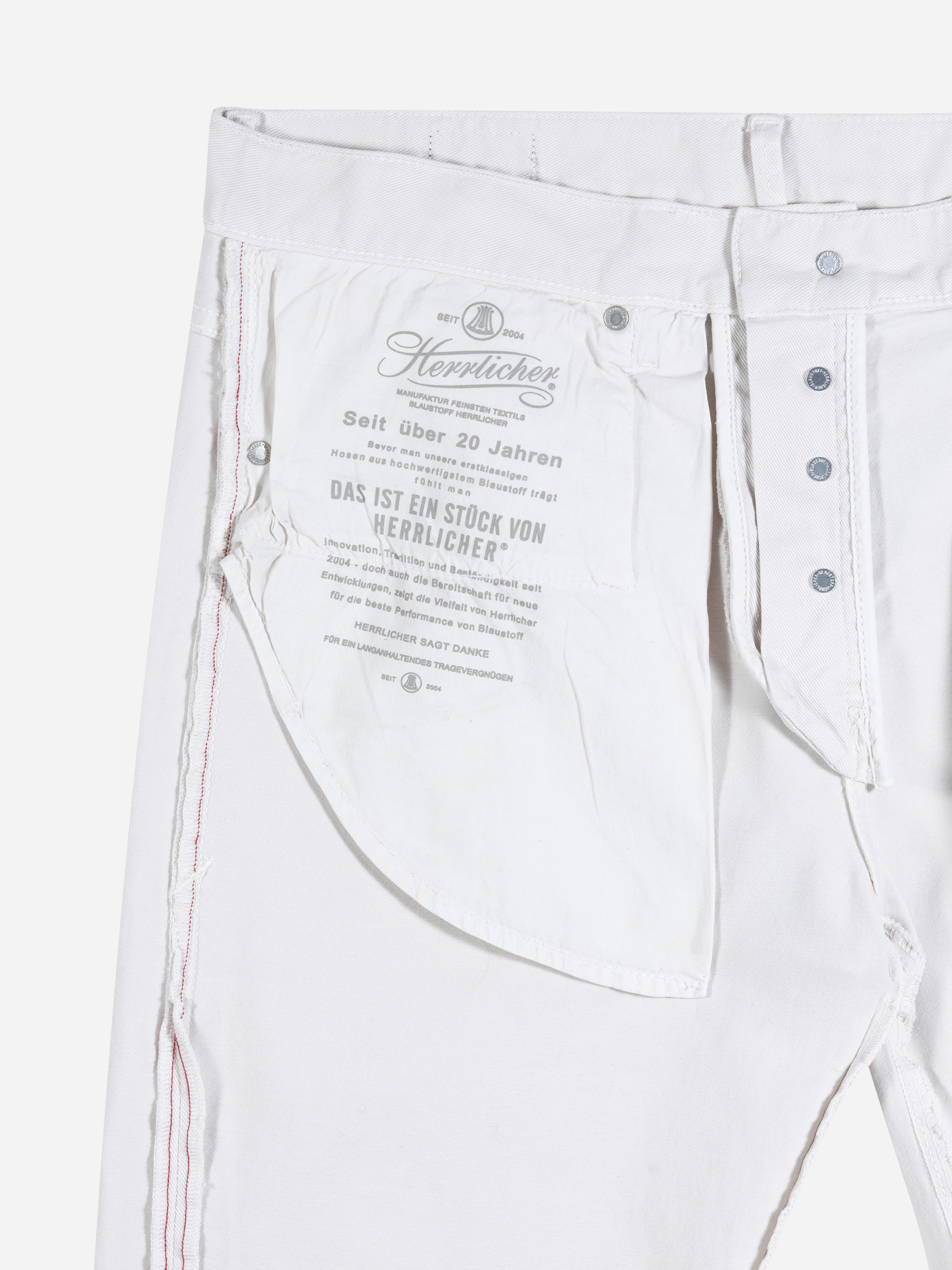 Weiße Denim-Jeans, bei der der Bund aufgeknöpft und die Tasche auf links gedreht ist, so dass der aufgedruckte Text in deutscher Sprache auf dem Taschenstoff zu sehen ist. Die Jeans hat einen sichtbaren Knopfverschluss und rote Nähte an der Naht.