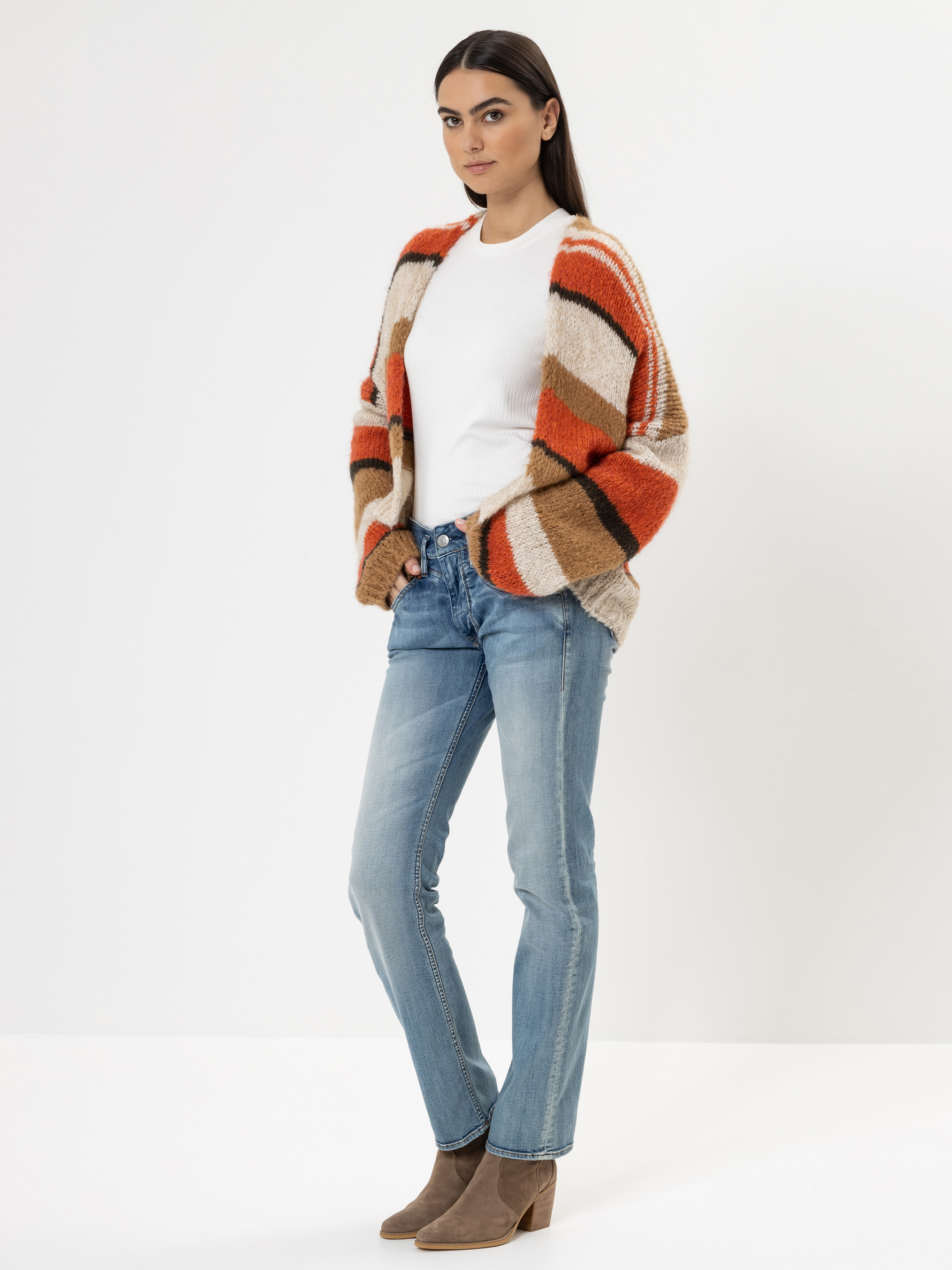 Eine Frau steht vor einem weißen Hintergrund und trägt ein weißes Hemd, blaue Jeans, taupefarbene Ankle Boots und eine gemütliche Strickjacke mit orangefarbenen, beigen und braunen Streifen. Sie hat langes dunkles Haar und einen neutralen Gesichtsausd