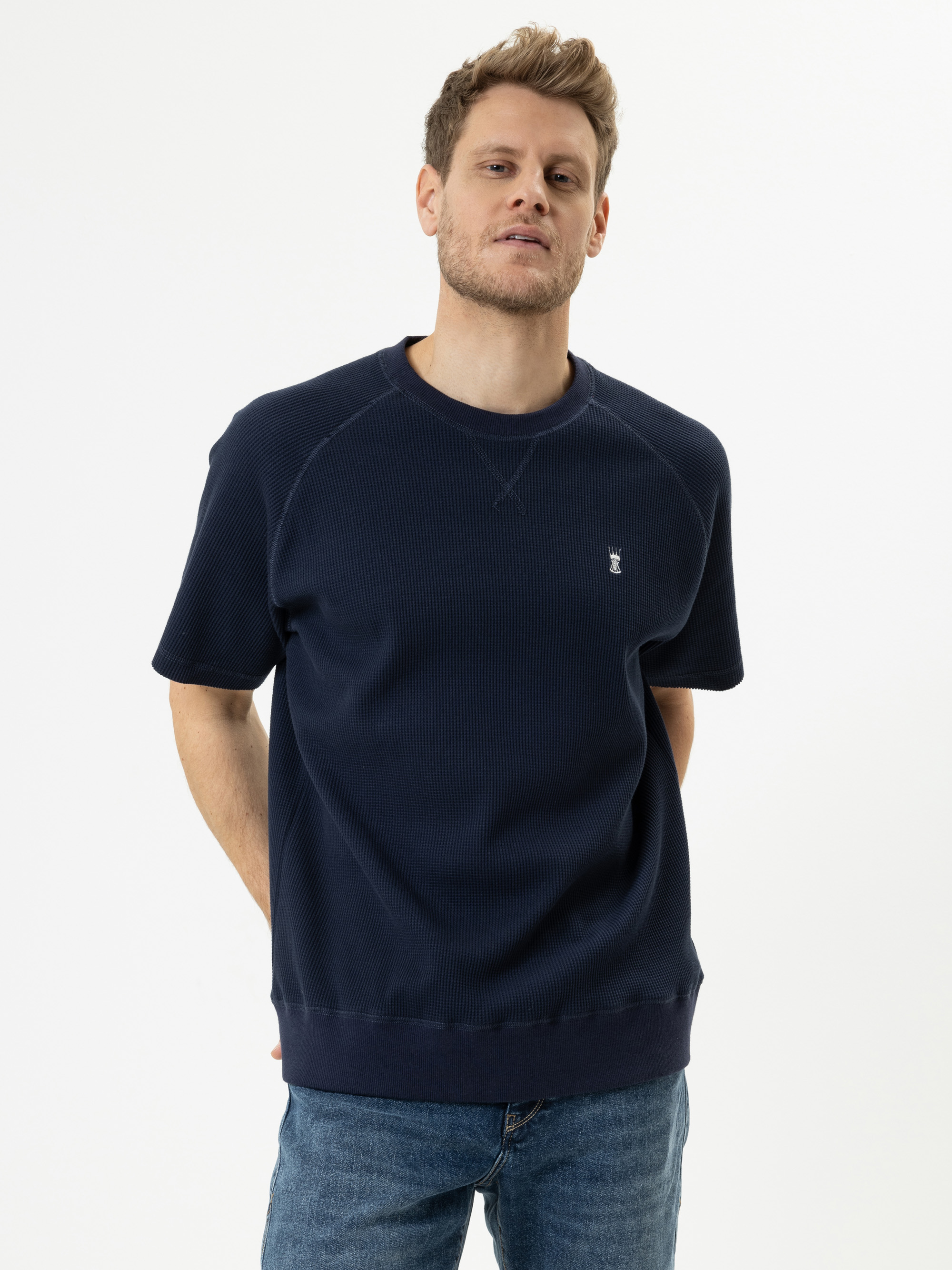 Ein Mann mit kurzen, hellbraunen Haaren, der ein marineblaues, kurzärmeliges Sweatshirt und blaue Jeans trägt, steht mit den Händen auf dem Rücken vor einem schlichten weißen Hintergrund.