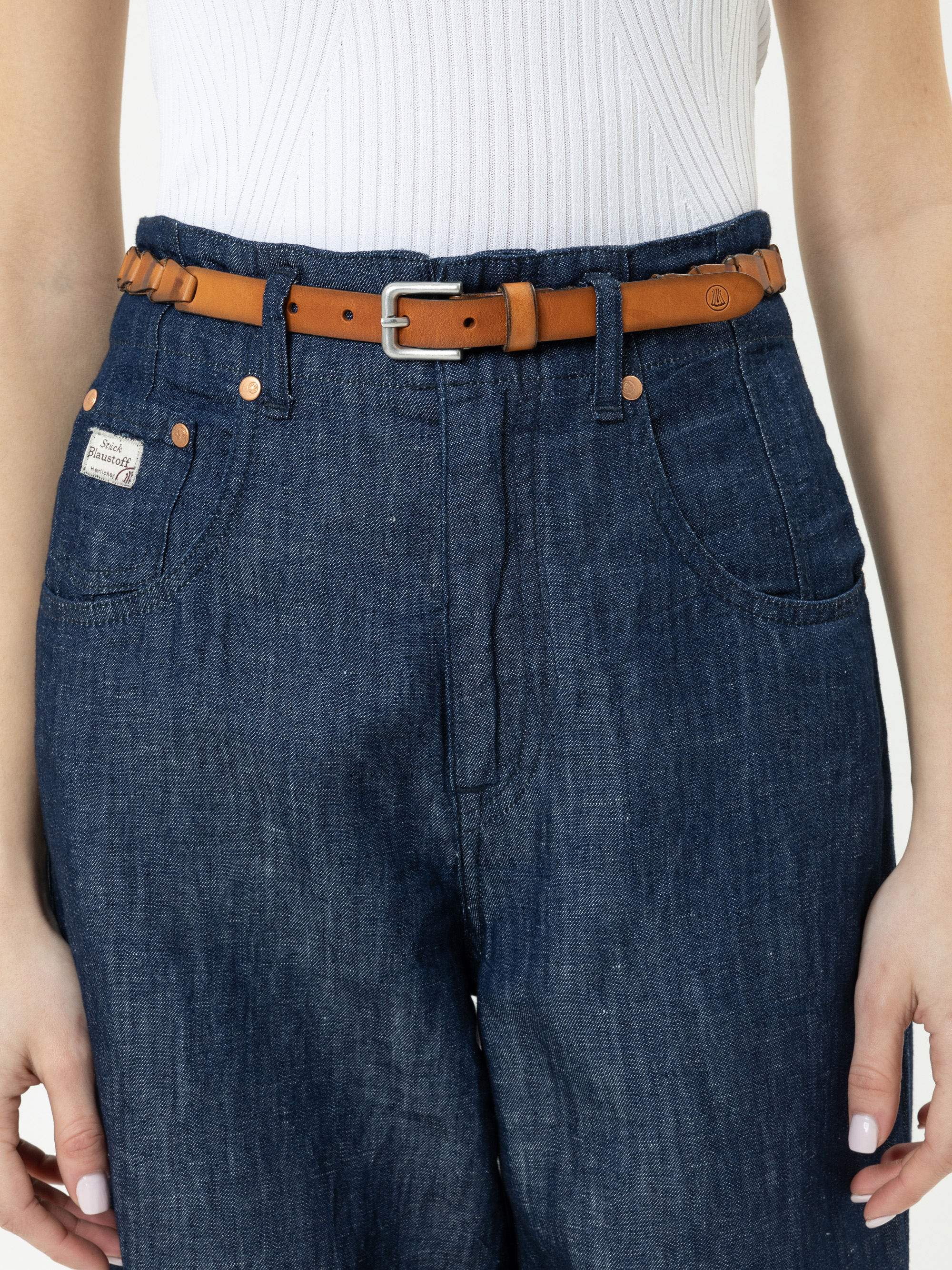 Eine Person, die eine blaue Jeans mit hoher Taille, einen braunen Ledergürtel und ein weißes geripptes Oberteil trägt, von der Taille bis zur Mitte des Oberschenkels, die Hände locker an der Seite.