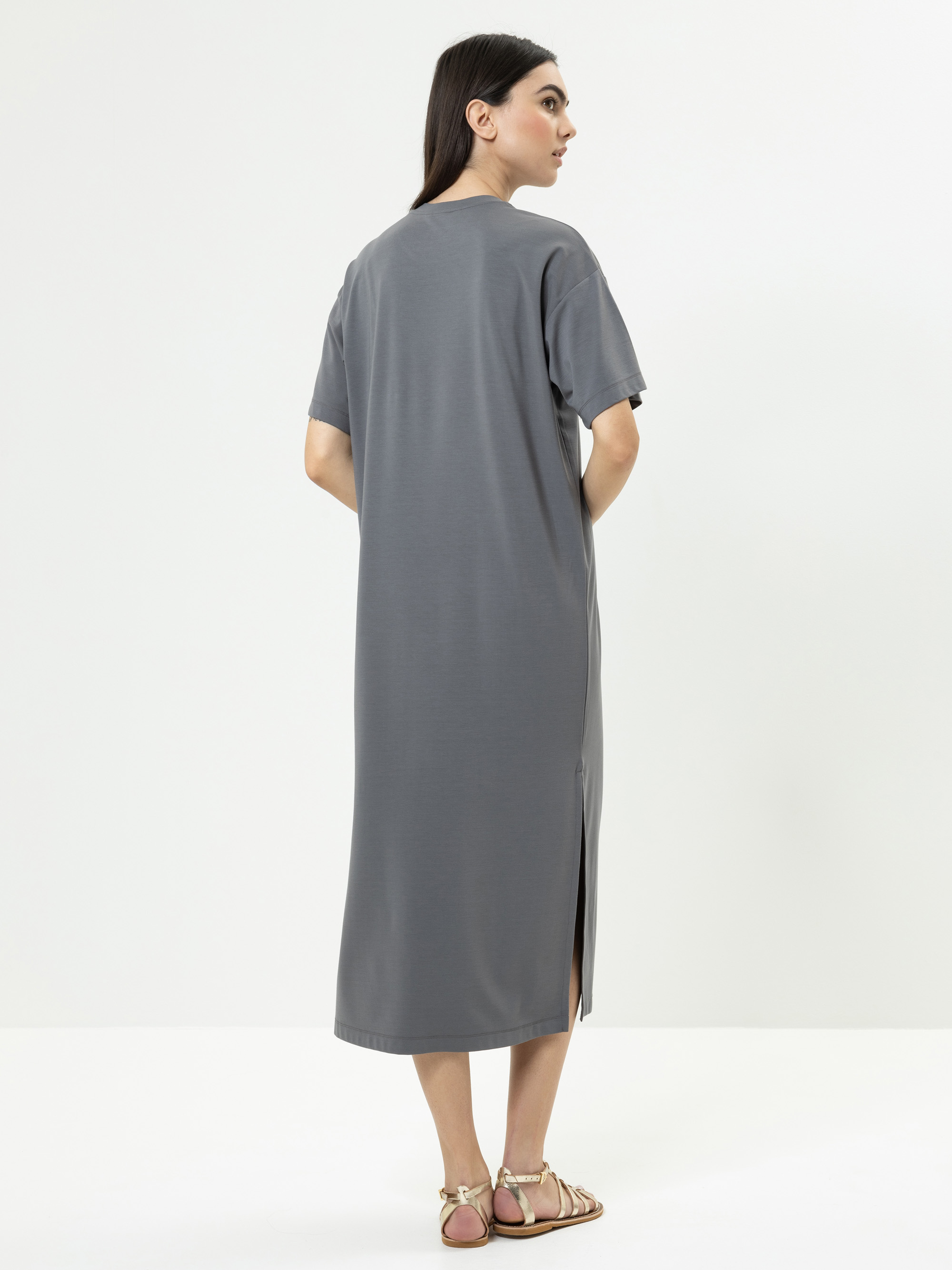 Eine Frau steht mit dem Gesicht zur Wand und trägt ein locker sitzendes, kurzärmeliges, graues Midikleid mit einem Seitenschlitz und flachen Sandalen. Sie befindet sich in einer minimalistischen, weißen Studioumgebung.