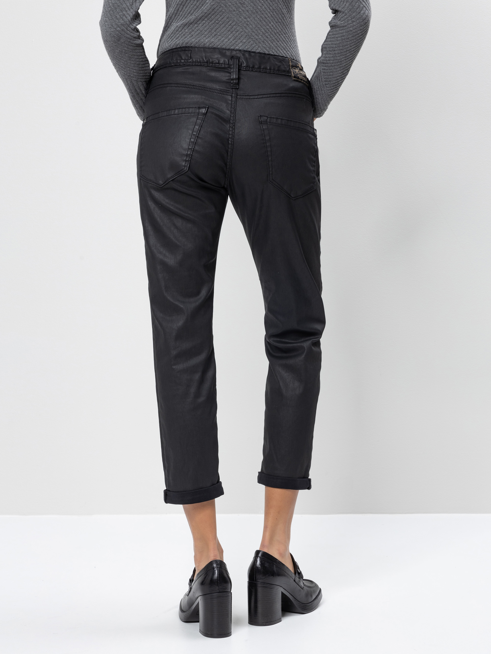 Eine Person, die schwarze Jeans mit Bündchen und schwarze Halbschuhe mit Absatz trägt, wird von hinten gezeigt, vor einem schlichten hellen Hintergrund. Die Person trägt außerdem ein langärmeliges, graues Oberteil.