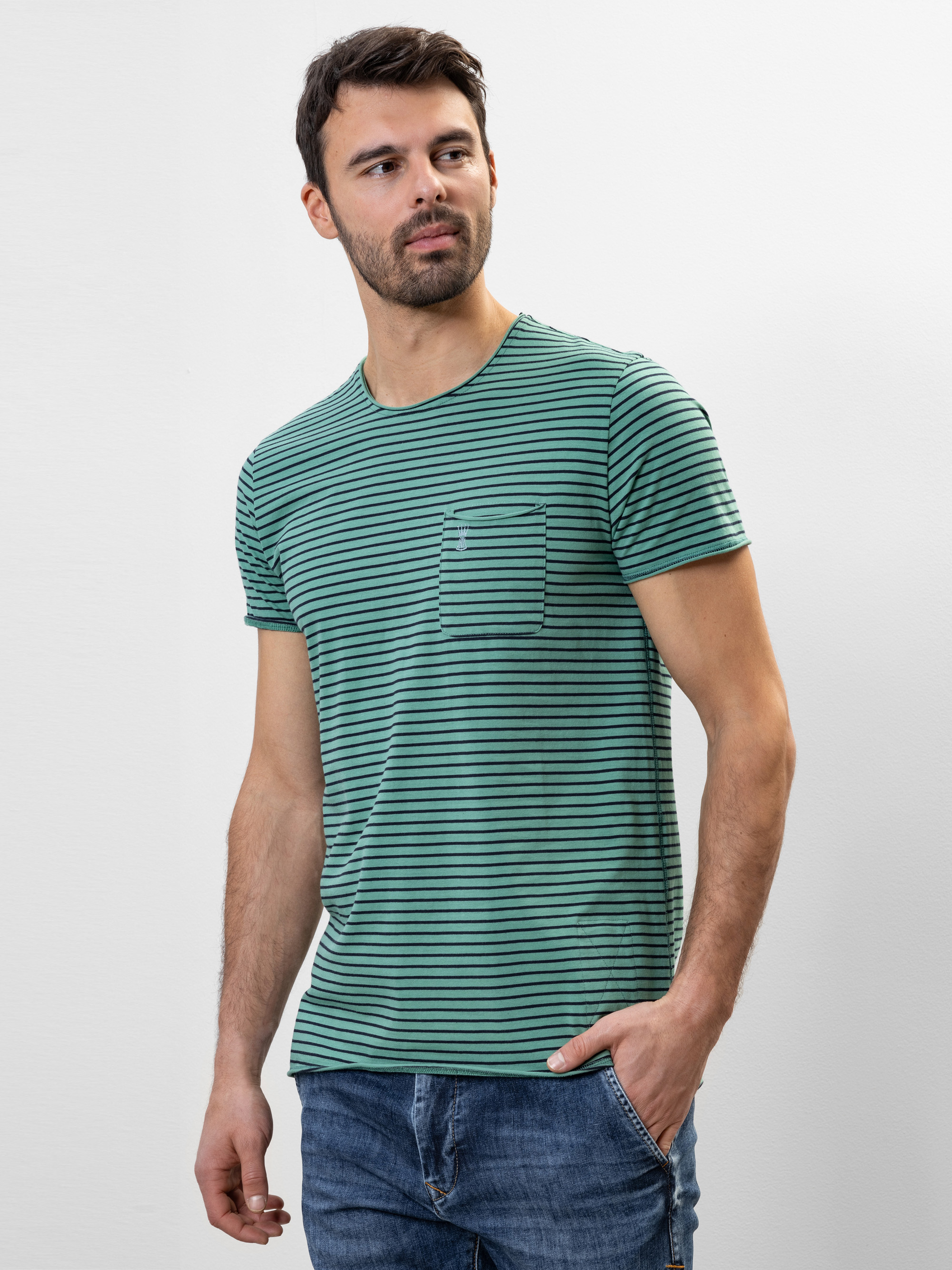 Ein Mann, der ein grün-schwarz gestreiftes kurzärmeliges T-Shirt mit einer kleinen Brusttasche und eine blaue Jeans trägt, steht vor einem schlichten weißen Hintergrund und schaut leicht zur Seite.