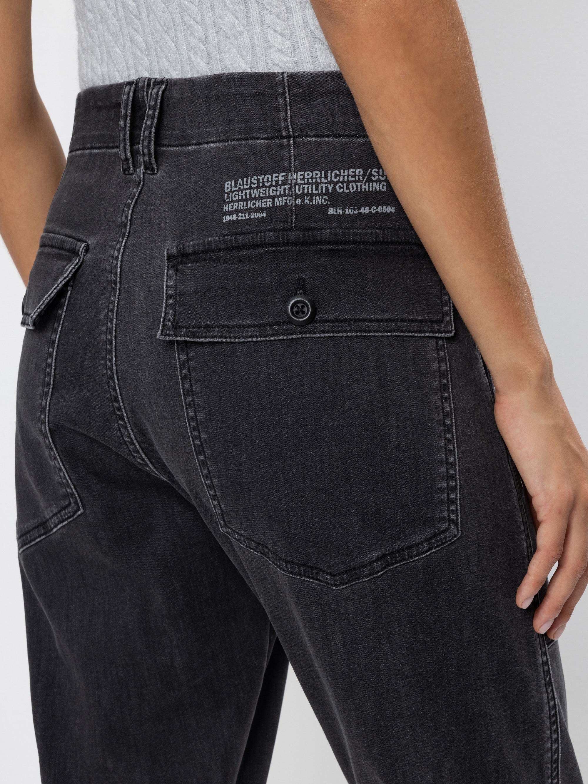Eine Person trägt eine dunkelgraue Utility-Jeans mit großen Gesäßtaschen und weißem Aufdruck über der rechten Tasche, von der Taille abwärts und von hinten gesehen.