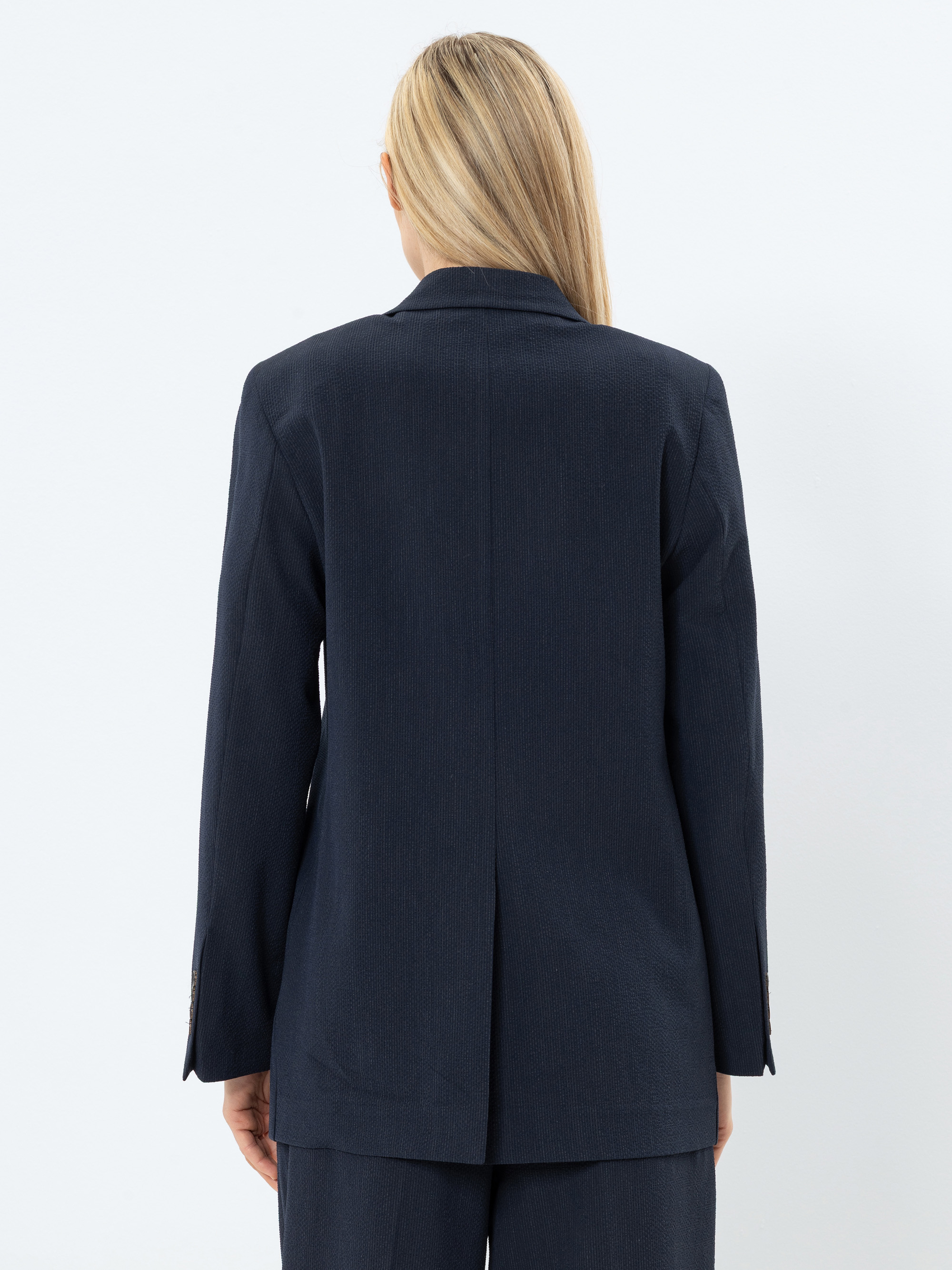 Eine Person mit langen blonden Haaren, von hinten gesehen, trägt einen dunklen marineblauen Blazer und eine passende Hose vor einem schlichten weißen Hintergrund.