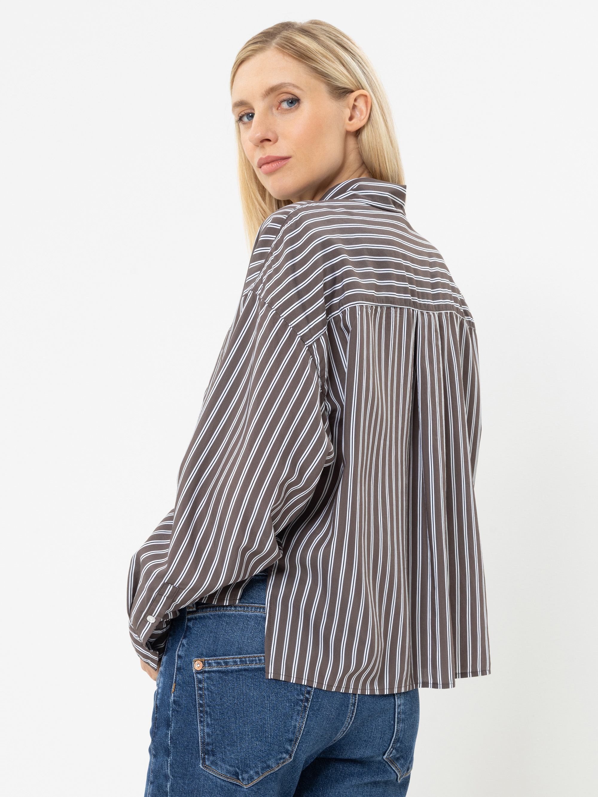 Eine Frau mit langen blonden Haaren, die ein braun-weiß vertikal gestreiftes Shirt und blaue Jeans trägt und vor einem schlichten weißen Hintergrund über die Schulter blickt.