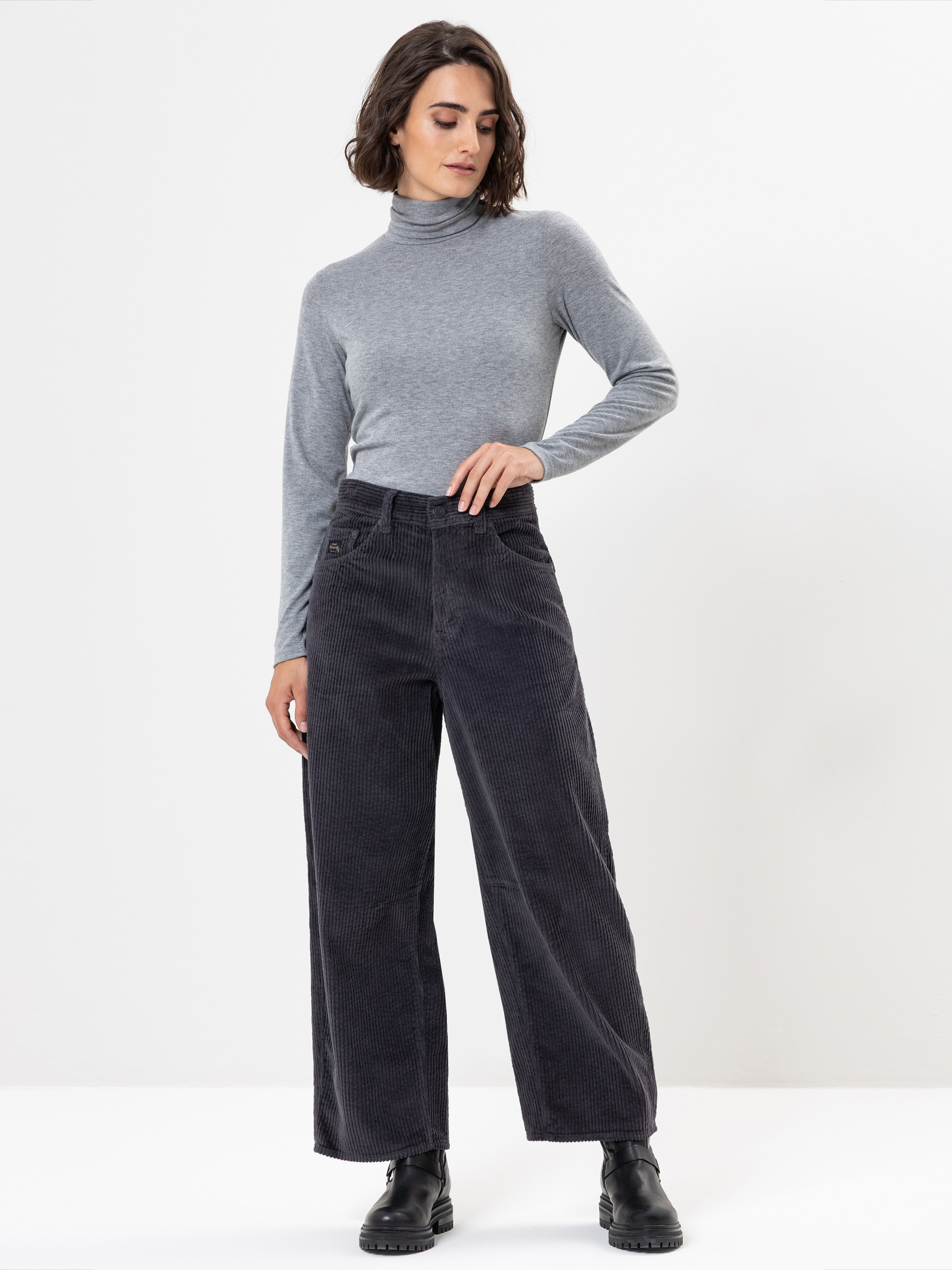 Eine Frau steht vor einem schlichten weißen Hintergrund und trägt ein hellgraues Rollkragenoberteil, eine dunkle, weit geschnittene Cordhose mit hoher Taille und schwarze Stiefel. Sie blickt leicht nach unten und hat eine Hand auf die Hüfte gestützt.