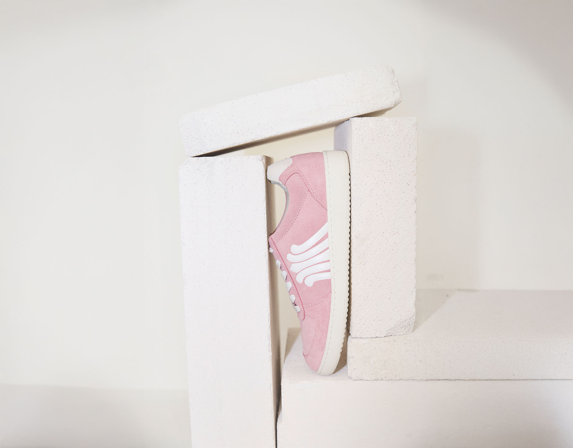 Ein rosafarbener Turnschuh mit weißen Akzenten steht aufrecht zwischen gestapelten weißen rechteckigen Blöcken und bildet ein minimalistisches und modernes Bild vor einem schlichten Hintergrund.
