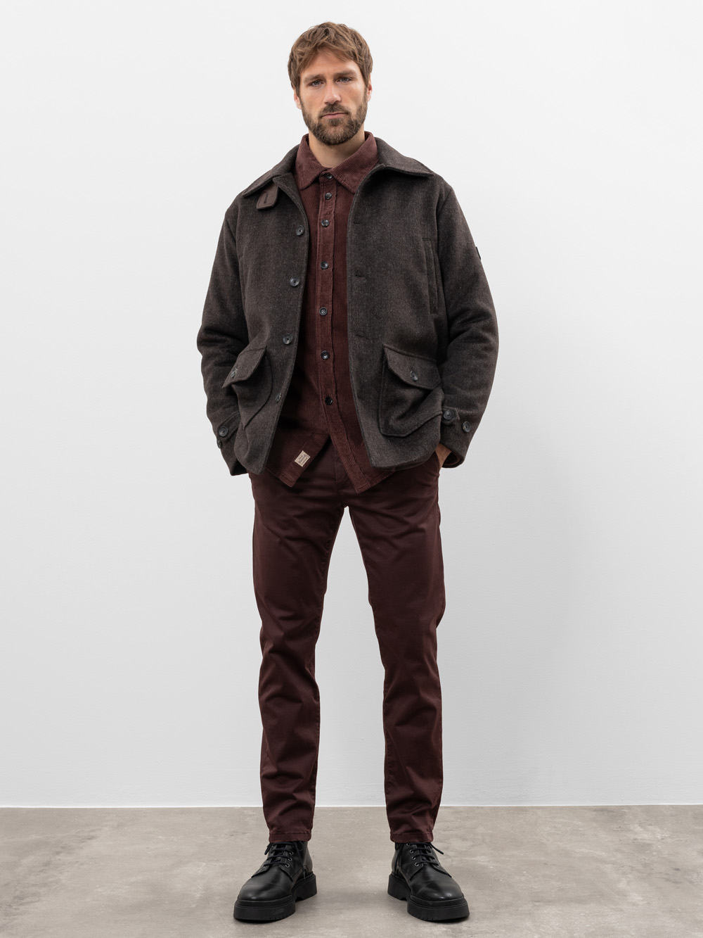 Ein Mann steht vor einer schlichten hellgrauen Wand und trägt eine dunkelbraune Jacke, ein kastanienbraunes Button-up-Hemd, eine kastanienbraune Hose und schwarze Schuhe. Seine Hände stecken in den Jackentaschen und er schaut direkt in die Kamera.