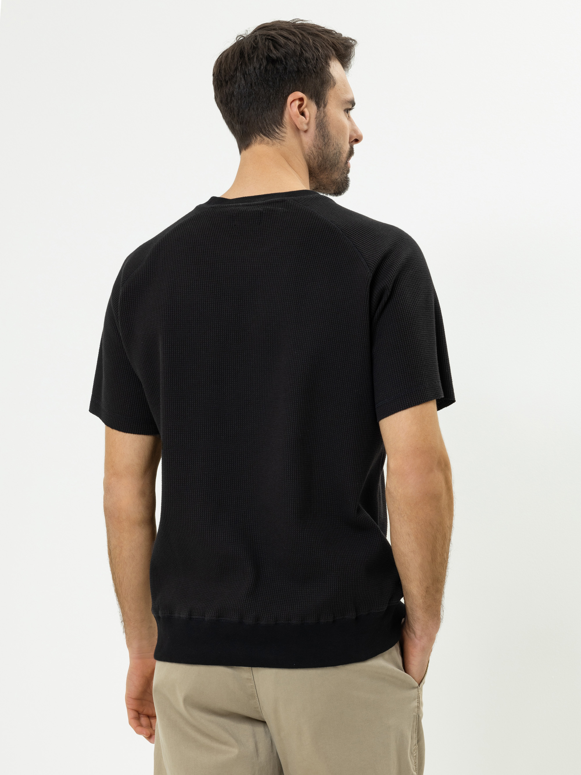 Ein Mann mit kurzen braunen Haaren, der ein schwarzes, kurzärmeliges Hemd und eine beigefarbene Hose trägt, steht mit dem Gesicht nach hinten und zeigt seinen Rücken vor einem schlichten weißen Hintergrund.