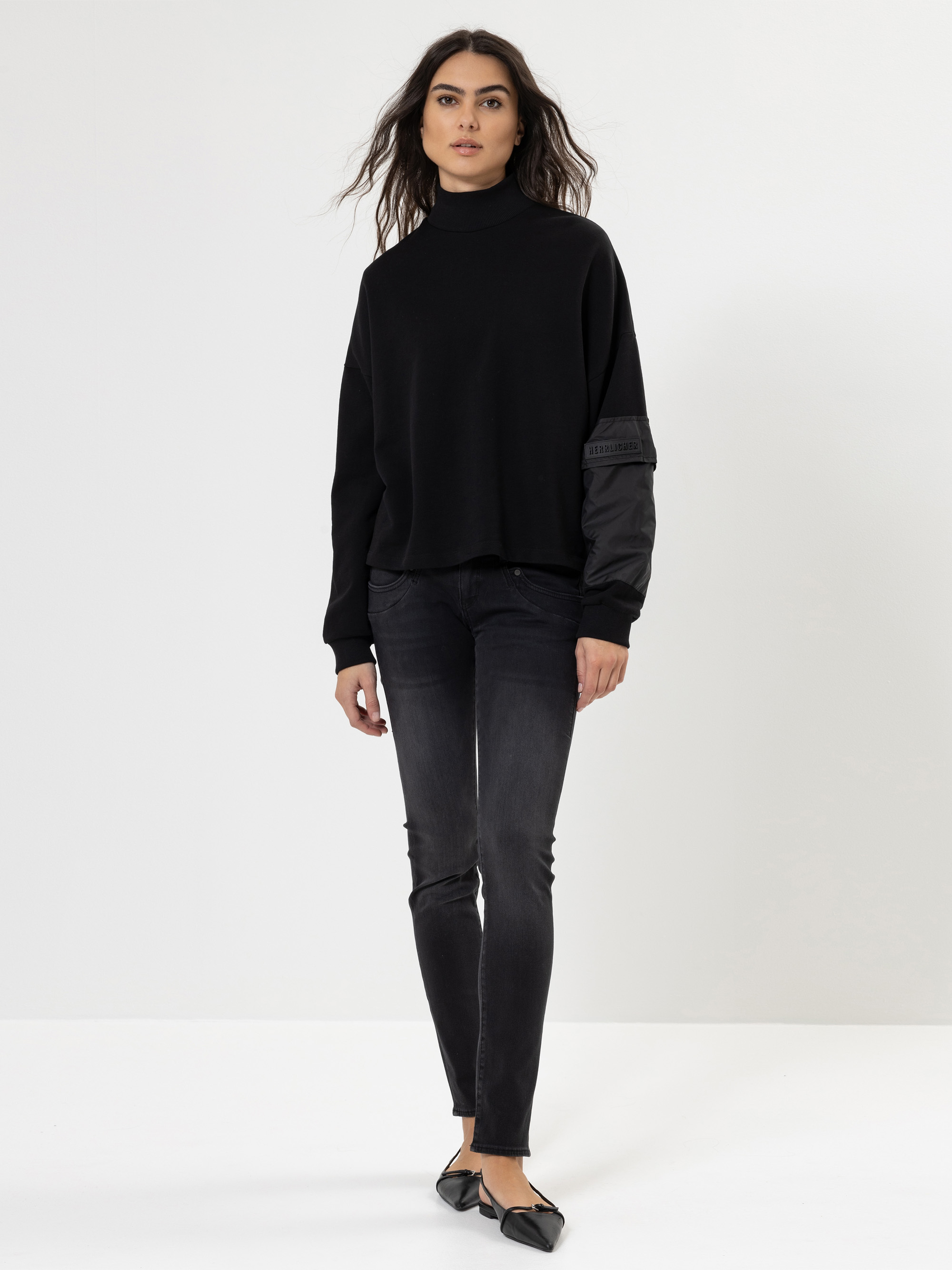 Eine Frau steht vor einem schlichten weißen Hintergrund und trägt einen schwarzen Rollkragenpullover, dunkle Röhrenjeans und schwarze spitze Schuhe. Sie hat langes gewelltes Haar und einen neutralen Gesichtsausdruck, eine Hand hält sie an der Seite.