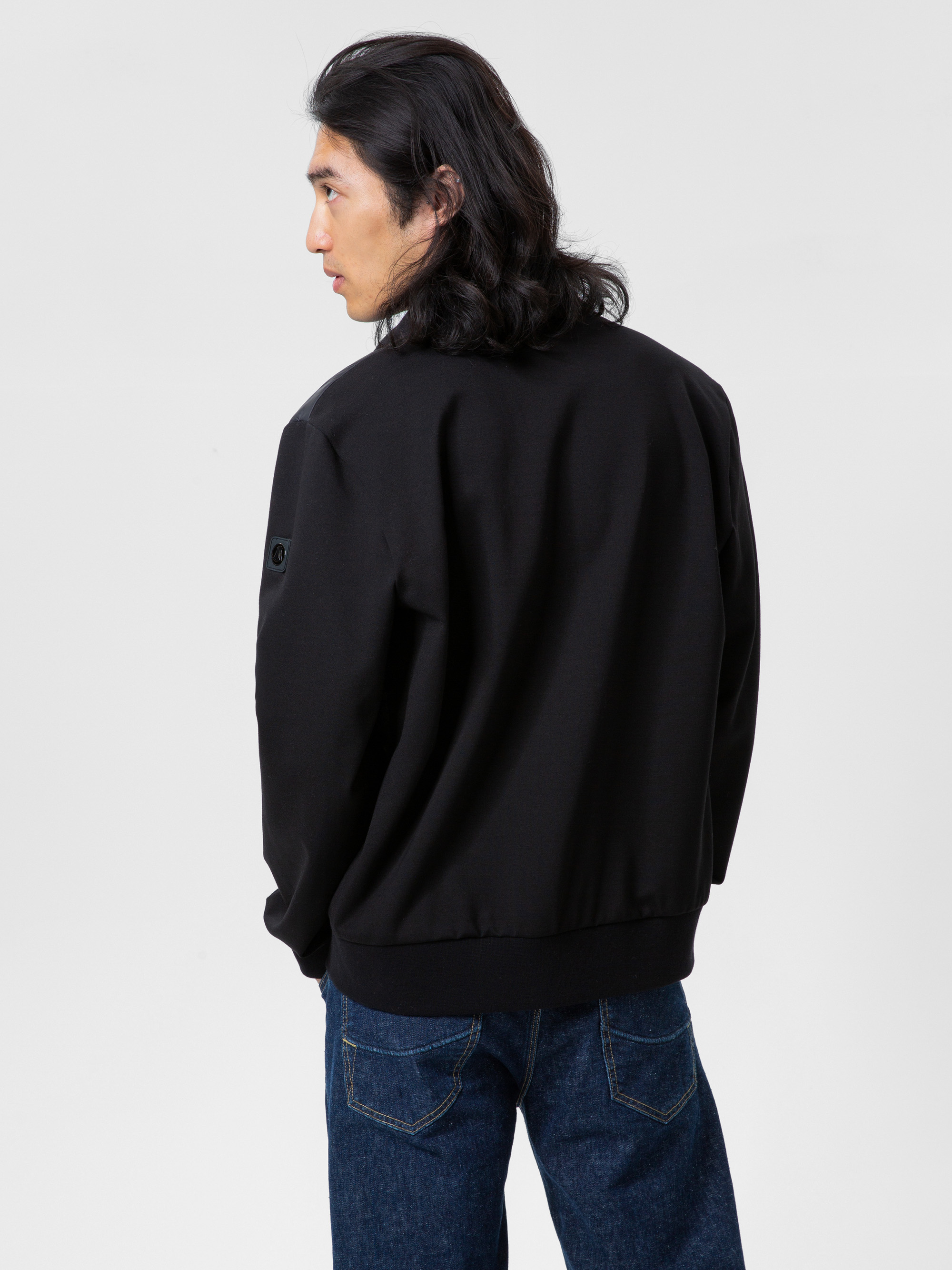 Ein Mann mit schulterlangen dunklen Haaren steht mit schwarzer Jacke und blauer Jeans vor einem schlichten weißen Hintergrund und blickt über seine linke Schulter.