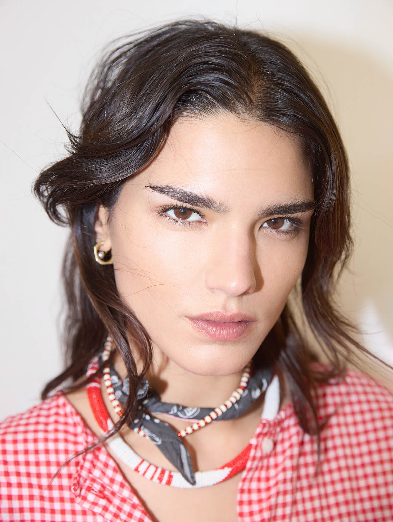 Eine Person mit langen dunklen Haaren, die ein rot-weiß kariertes Oberteil, Reifohrringe und mehrlagige Perlen- und Stoffketten trägt, blickt mit neutralem Blick direkt in die Kamera.