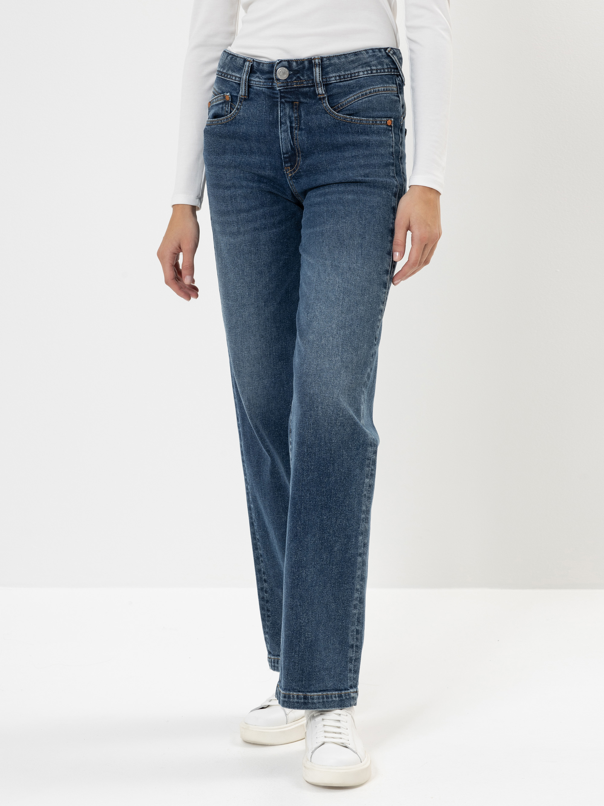 Eine Person, die eine blaue Jeans mit hoher Taille und geradem Bein, ein langärmeliges weißes Oberteil und weiße Turnschuhe trägt, steht vor einem schlichten weißen Hintergrund. Das Bild ist an den Schultern abgeschnitten.