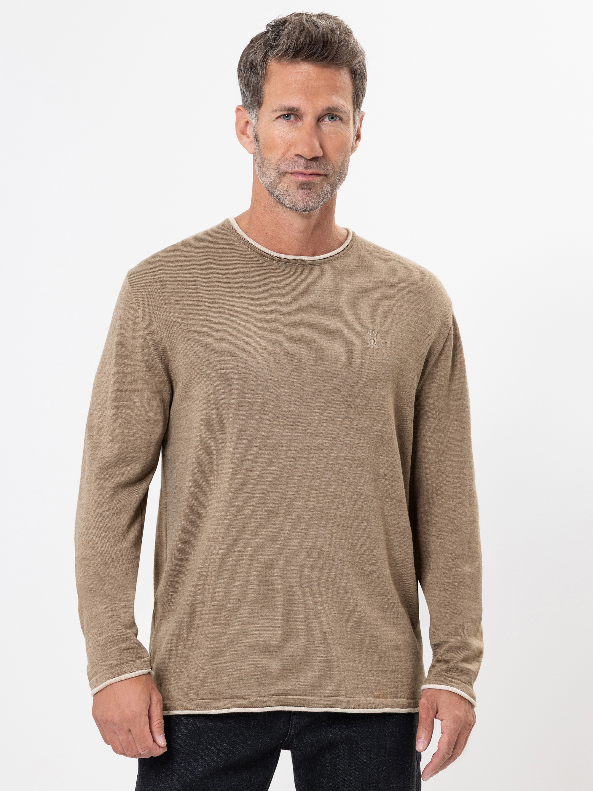 Ein Mann mit kurzen braunen Haaren und einem Bart trägt einen hellbraunen, langärmeligen Pullover mit Rundhalsausschnitt und weißem Rand und steht vor einem schlichten weißen Hintergrund.