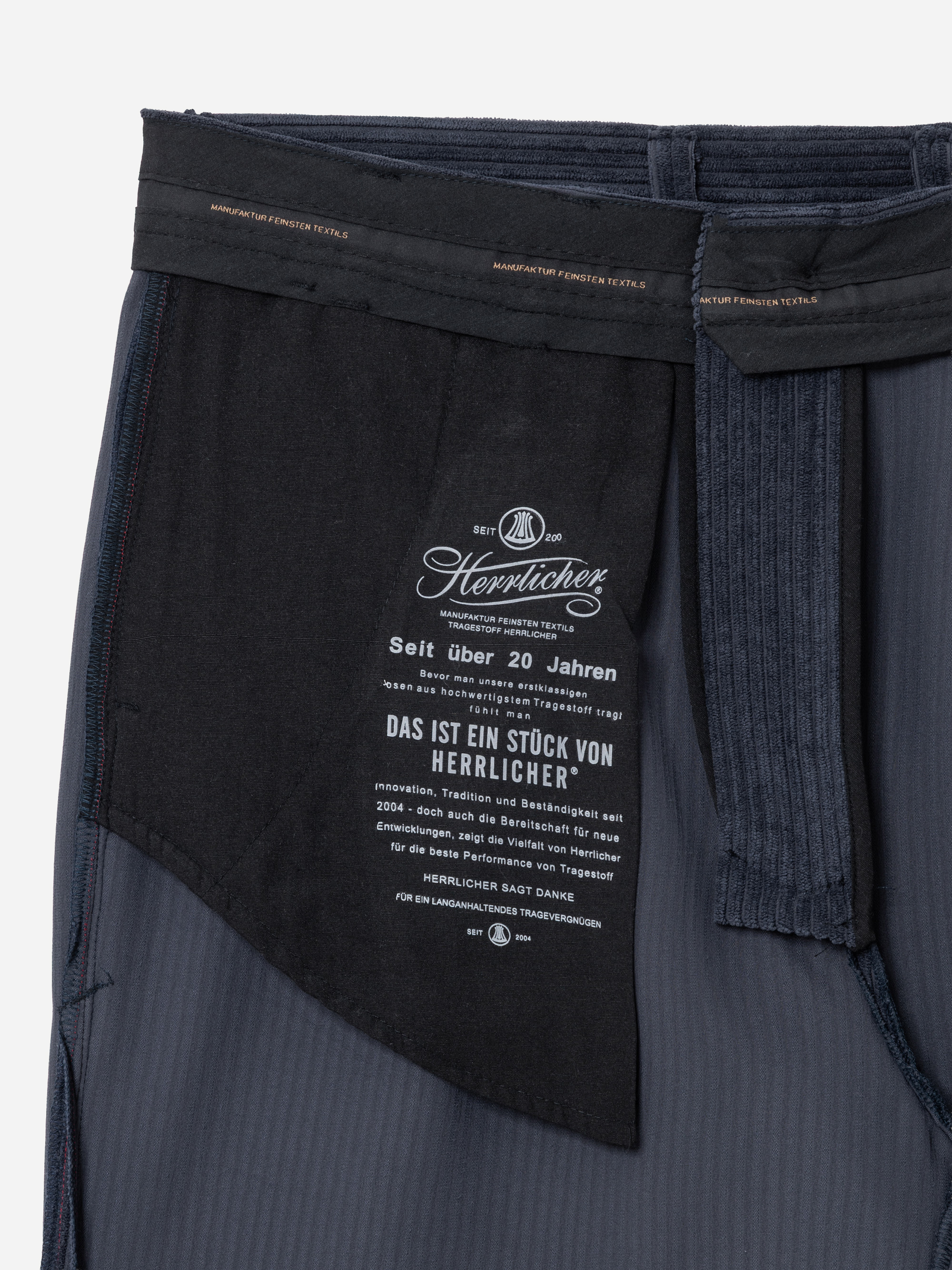 Nahaufnahme des Innenbereichs der Tasche einer dunkelblauen Herrlicher-Hose, die ein Stoffetikett mit weißem Text in deutscher Sprache, das Markenlogo sowie auf das Taschenfutter aufgedruckte Produktdetails zeigt.