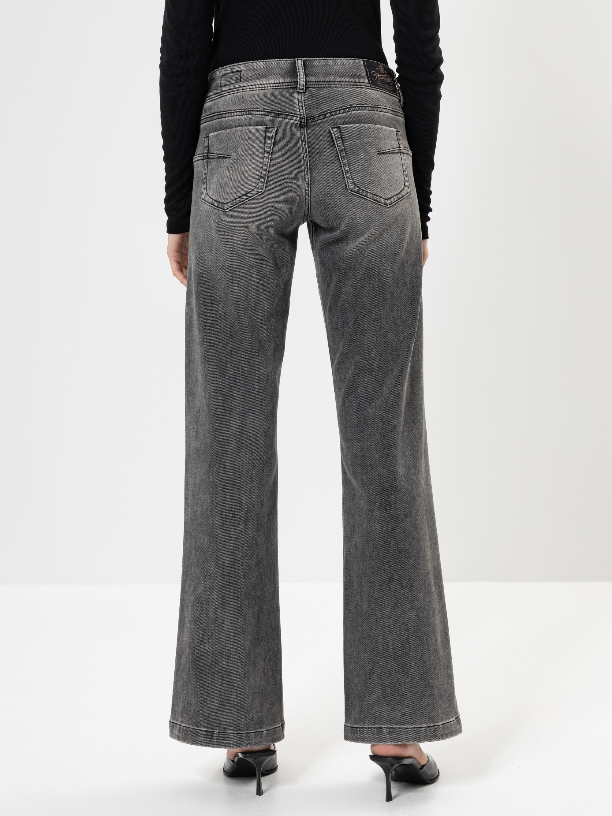 Eine Person, die eine graue Jeans mit hoher Taille und weitem Bein sowie schwarze Schuhe mit Absatz trägt, wird von hinten vor einem schlichten weißen Hintergrund gezeigt.