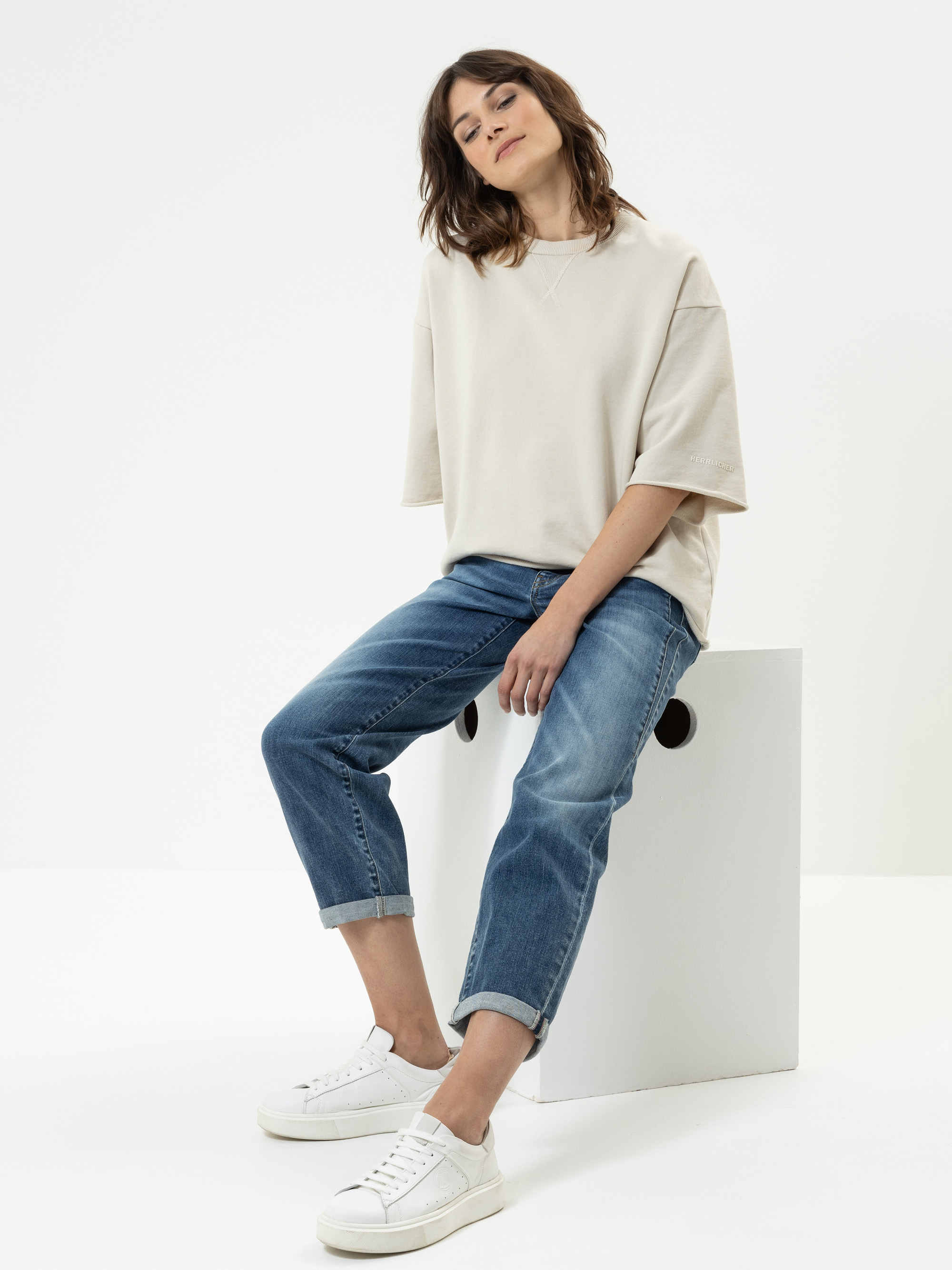 Eine Frau mit schulterlangen braunen Haaren sitzt auf einem weißen Block. Sie trägt ein beiges Oversized-T-Shirt, blaue Jeans mit Bündchen und weiße Turnschuhe vor einem schlichten weißen Hintergrund.