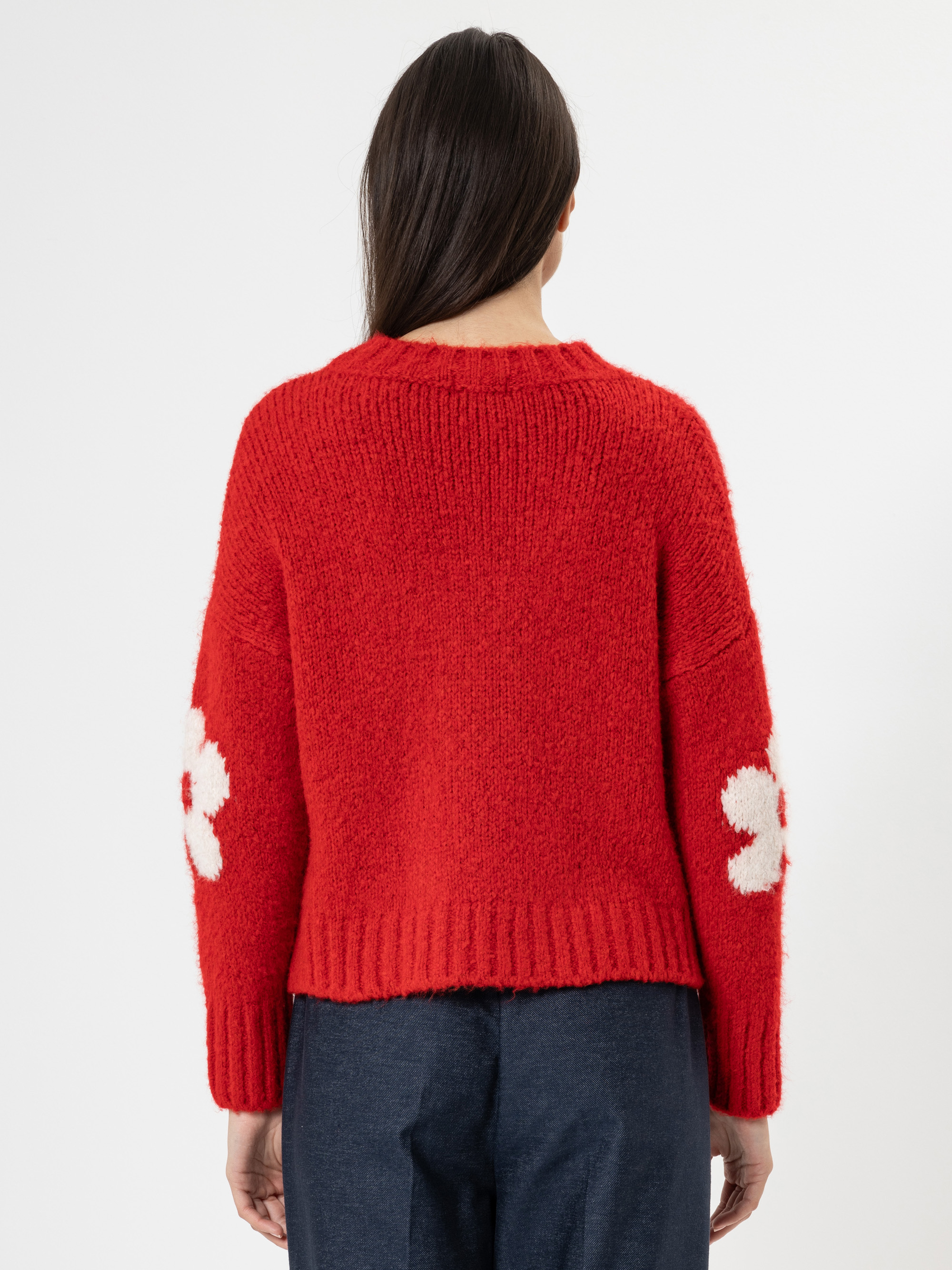 Eine Person mit langen dunklen Haaren, von hinten gesehen, trägt einen roten wuscheligen Pullover mit weißen Blumenmustern an den Ärmeln und eine dunkelblaue Hose. Der Hintergrund ist einfarbig weiß.