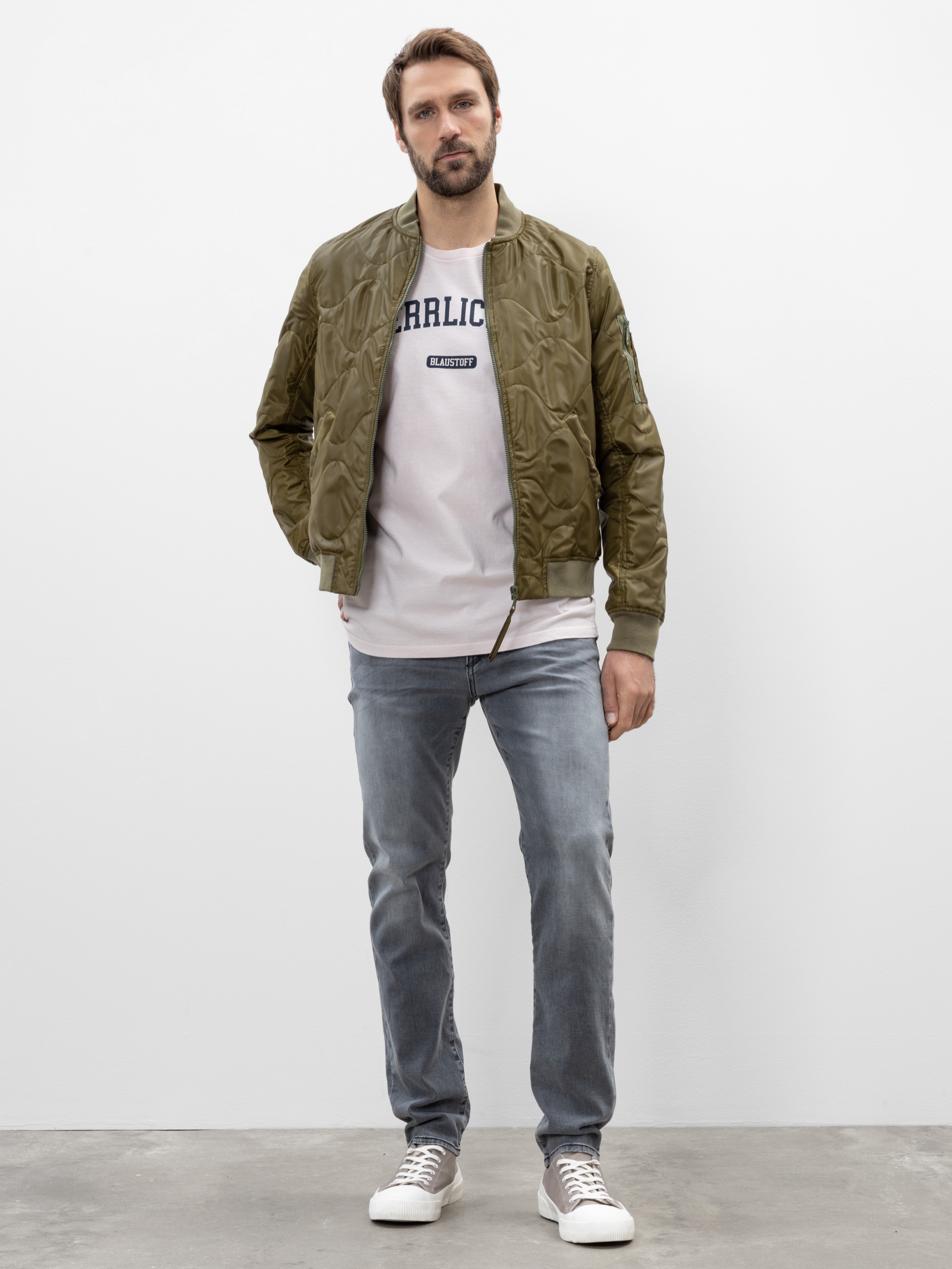 Ein Mann steht vor einer schlichten weißen Wand und trägt eine hellolivfarbene Bomberjacke, ein weißes grafisches T-Shirt, hellblaue Jeans und weiße Turnschuhe. Er hat eine Hand in der Tasche und einen neutralen Gesichtsausdruck.