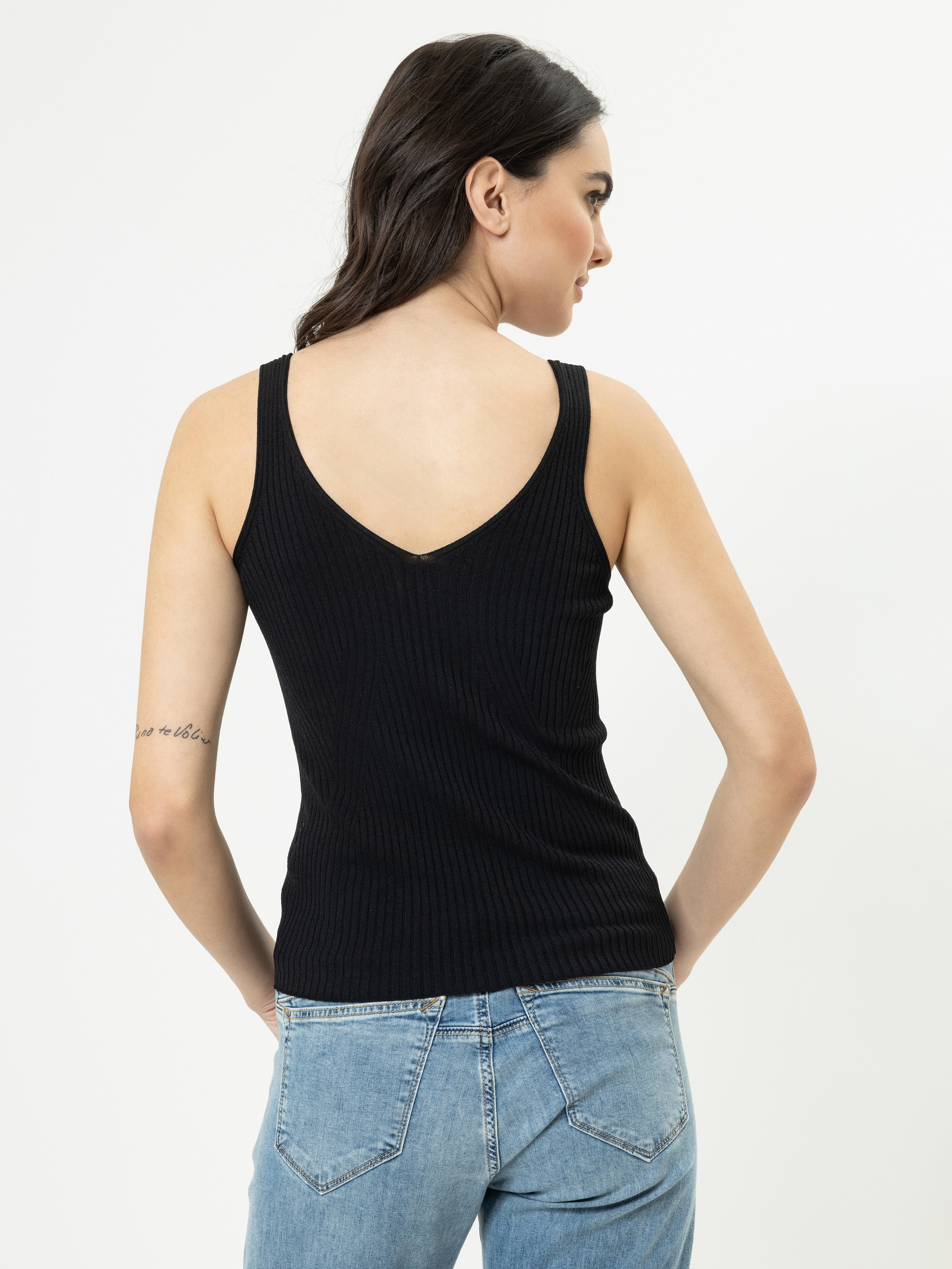 Eine Frau mit langen braunen Haaren, die ein schwarzes geripptes ärmelloses Oberteil und blaue Jeans trägt, steht mit dem Rücken zur Kamera vor einem schlichten weißen Hintergrund.