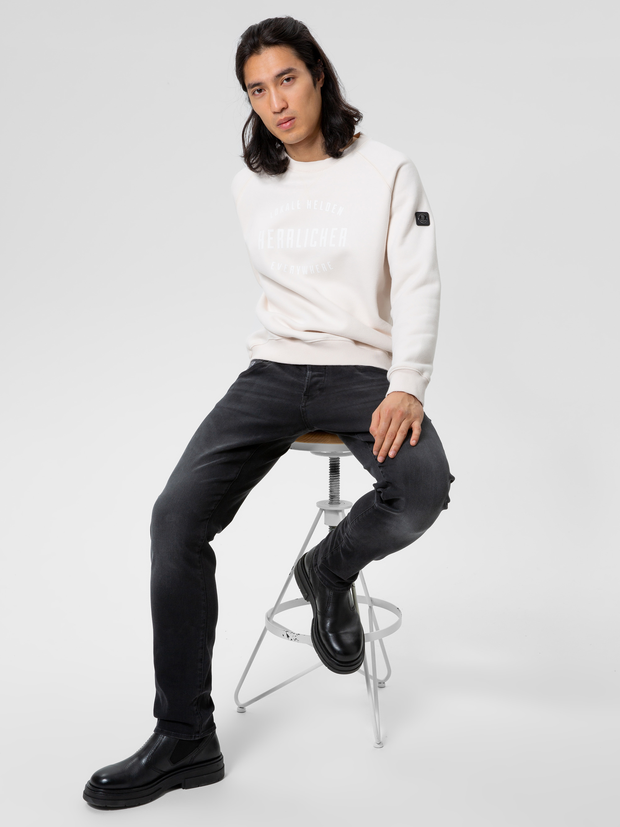 Eine Person mit langen dunklen Haaren, die ein helles Sweatshirt, schwarze Jeans und schwarze Stiefel trägt, sitzt auf einem hohen Hocker vor einem schlichten weißen Hintergrund.