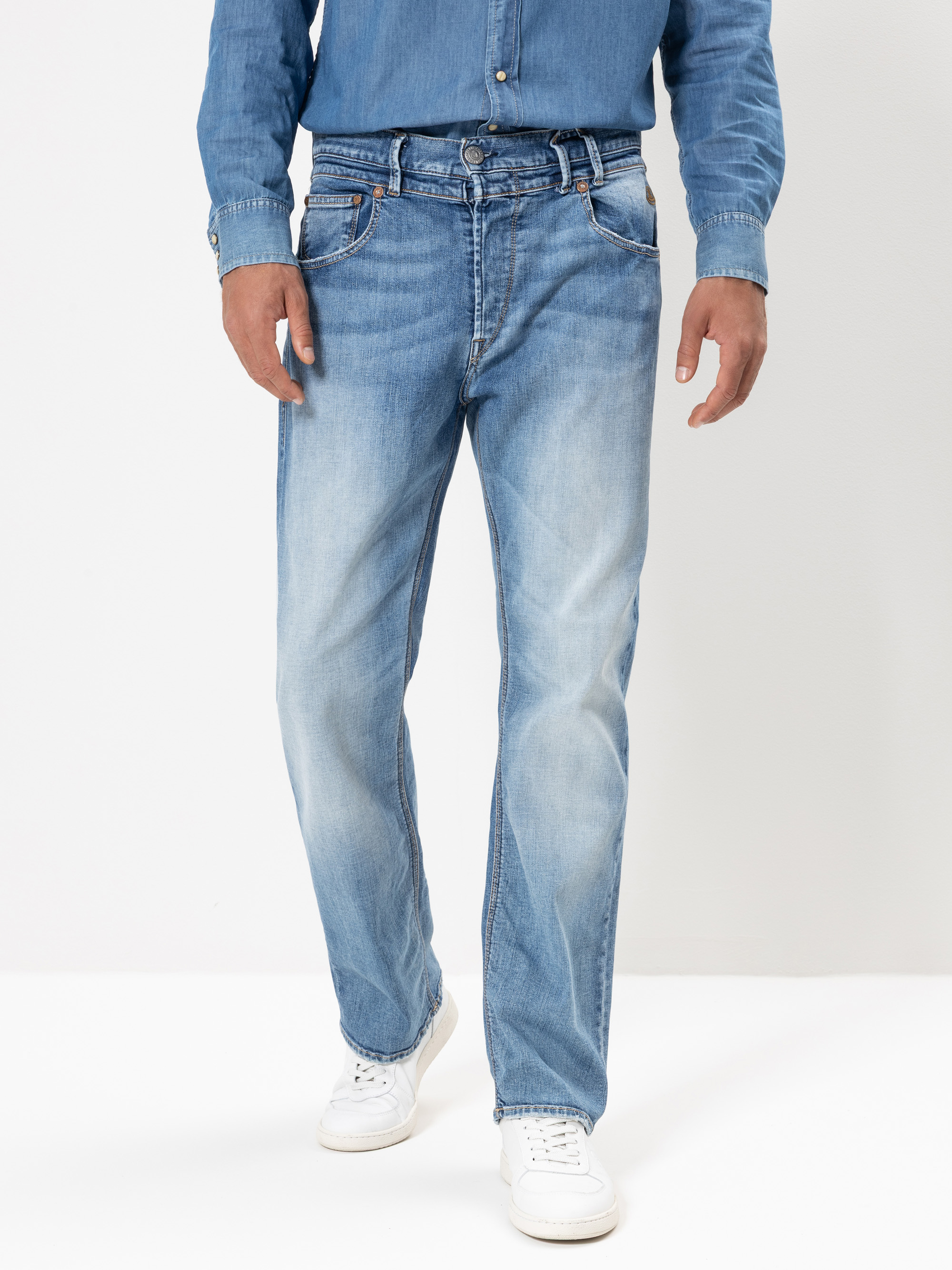 Ein Mann, der ein hellblaues Jeanshemd und hellblaue Jeans mit verblasster Waschung trägt, gepaart mit weißen Turnschuhen, steht vor einem schlichten weißen Hintergrund.