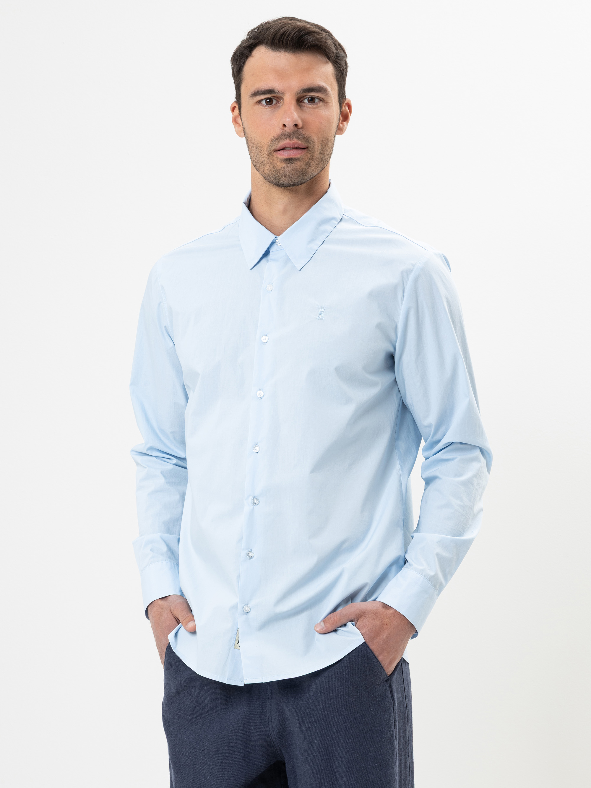 Ein Mann, der ein hellblaues Hemd mit Knopfleiste und eine dunkle Hose trägt, steht vor einem schlichten weißen Hintergrund, hat die Hände in den Taschen und schaut direkt in die Kamera.