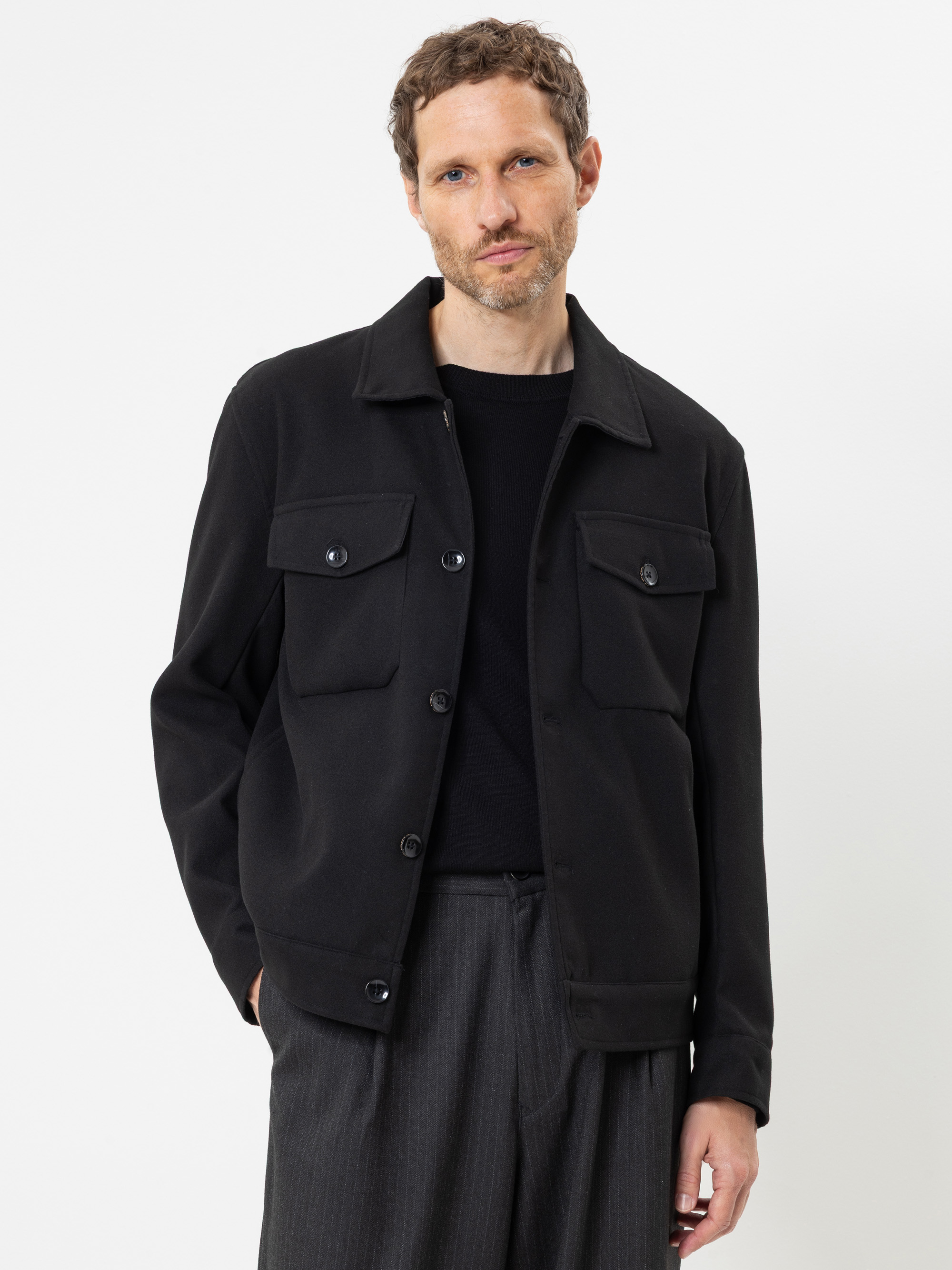 Ein Mann mit kurzen braunen Haaren und Bart trägt eine schwarze Jacke mit Knöpfen und Vordertaschen über einem schwarzen Hemd und einer grauen Hose und steht vor einem schlichten weißen Hintergrund.