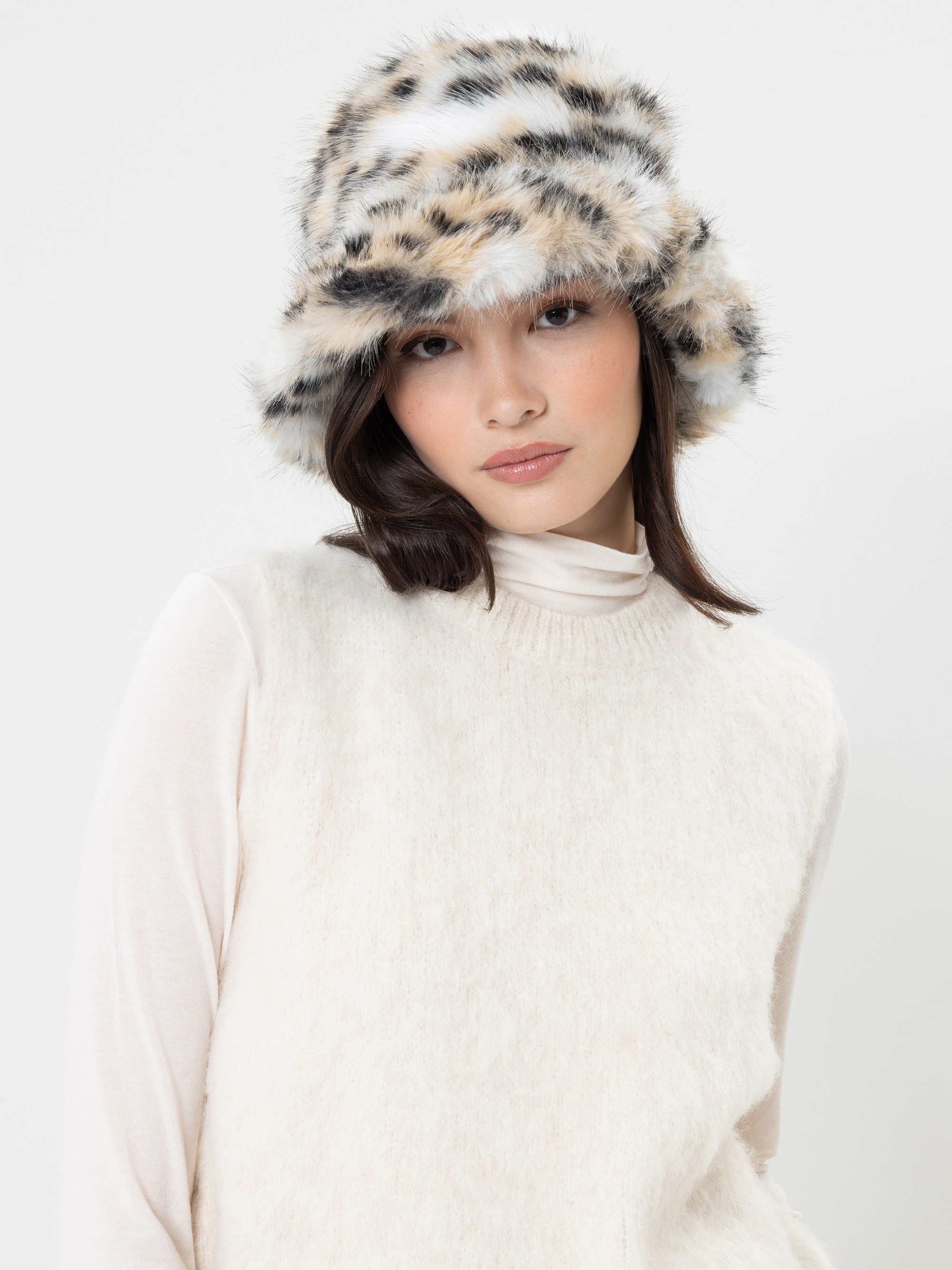Eine Frau, die einen cremefarbenen, ärmellosen Pullover über einem weißen Rollkragenpullover und einen flauschigen Bucket-Hat mit einem schwarz-beige-weißen Tiermuster trägt, steht vor einem schlichten weißen Hintergrund.