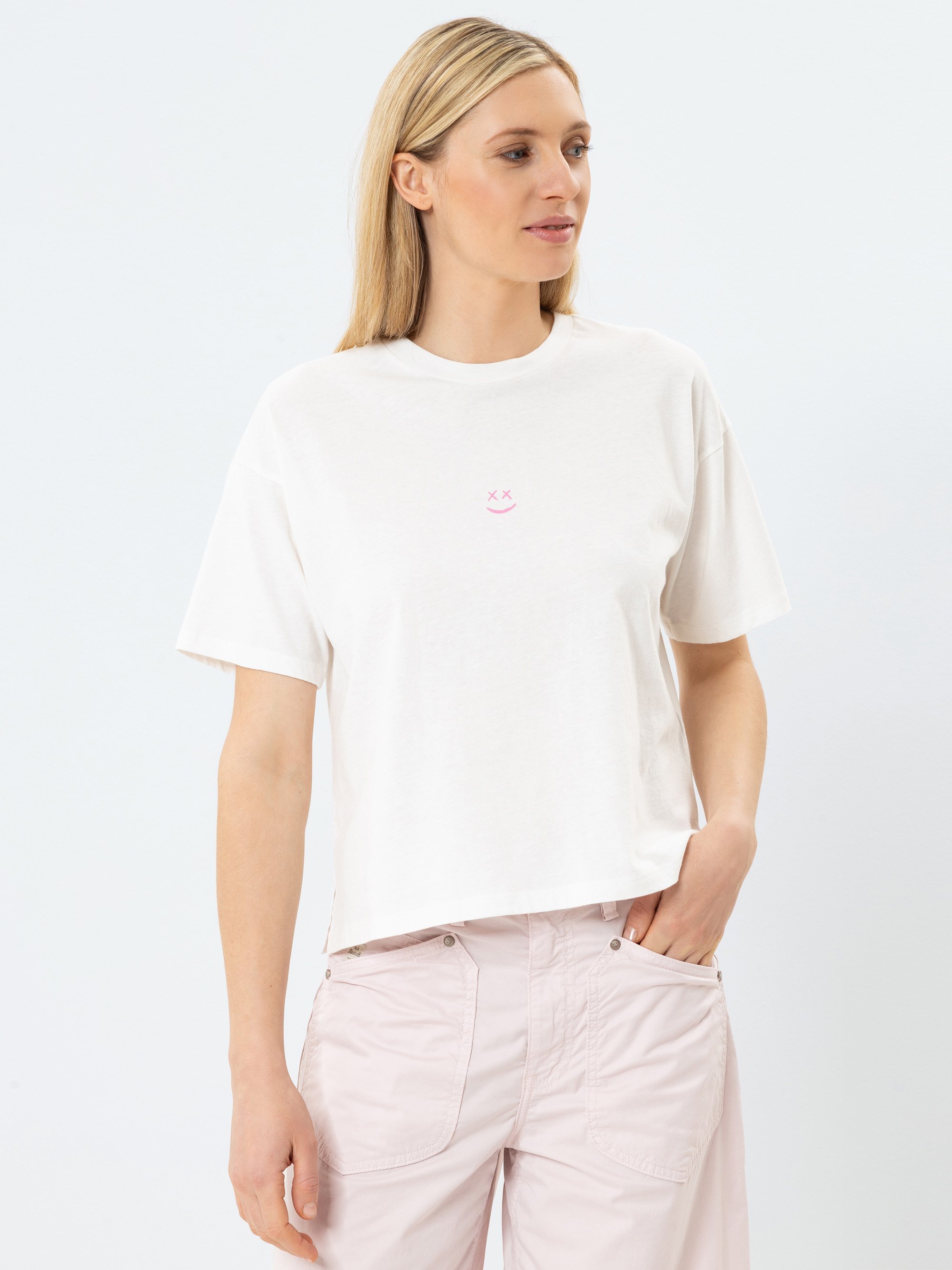 Eine Frau mit langen blonden Haaren trägt ein locker sitzendes weißes T-Shirt mit einem kleinen rosafarbenen Smiley-Motiv und eine hellrosafarbene Hose und steht vor einem schlichten hellen Hintergrund.