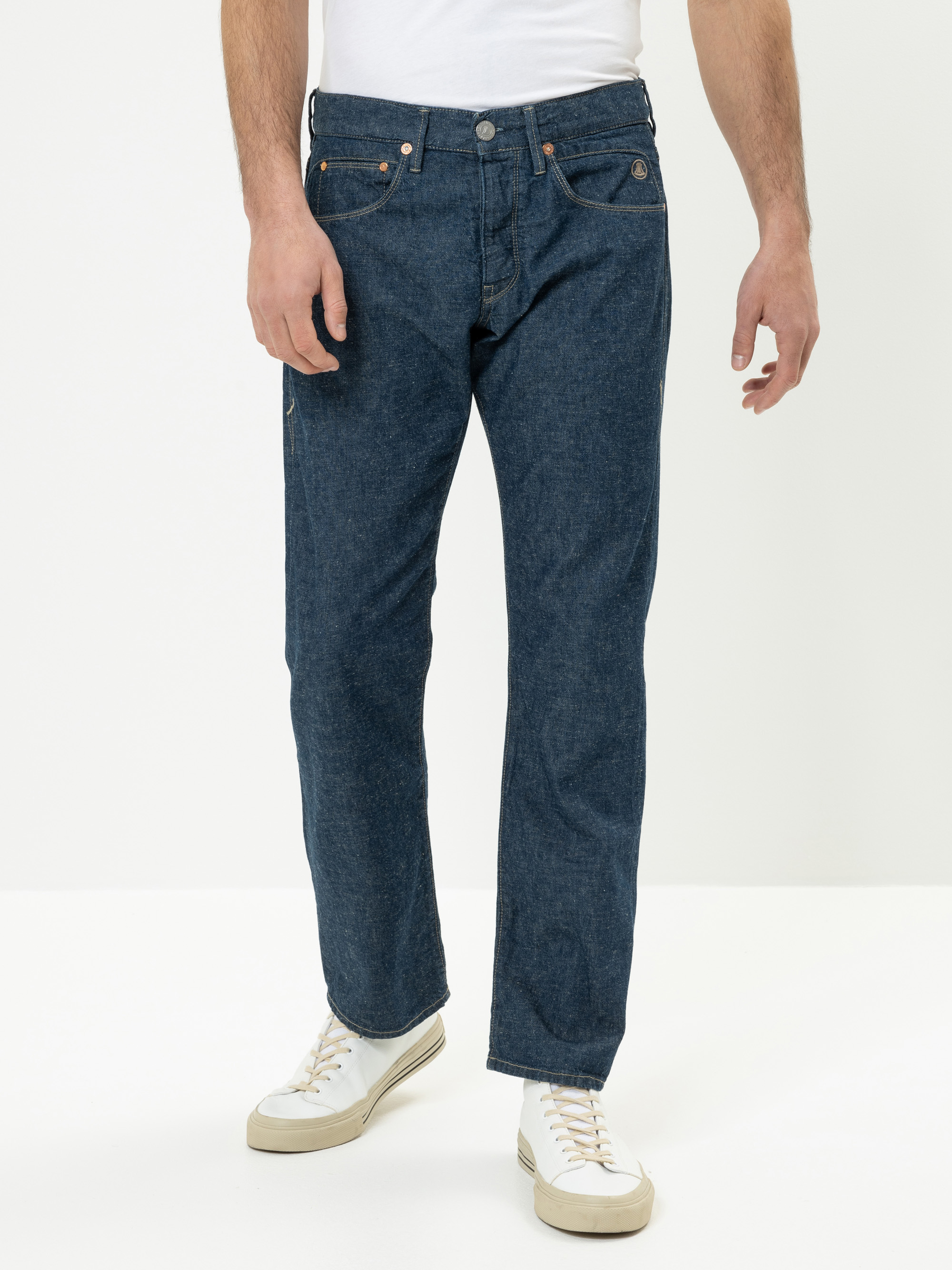 Eine Person, die blaue Jeans mit geradem Bein und weiße Turnschuhe trägt, steht vor einem schlichten weißen Hintergrund. Der Oberkörper der Person ist teilweise sichtbar, und die Arme liegen entspannt an der Seite.