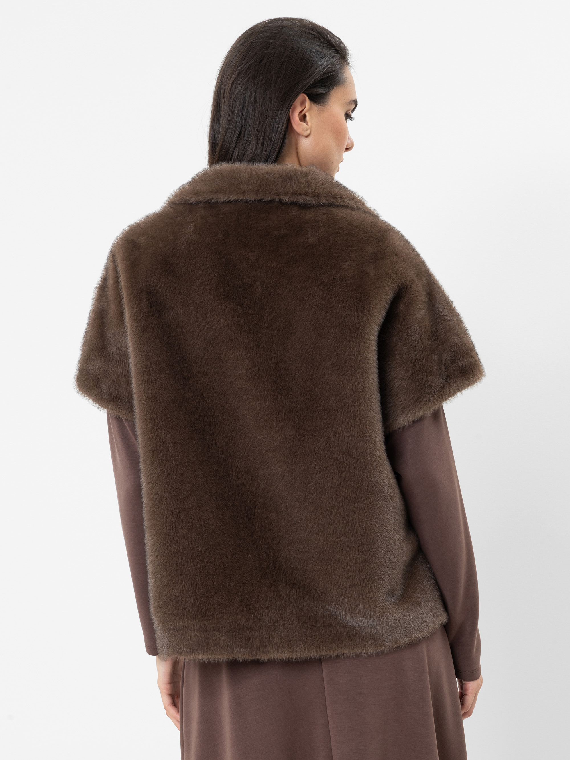 Eine Frau mit dunklem Haar, die eine braune Kurzarmjacke aus Kunstfell über einem braunen Langarmkleid trägt, steht mit dem Rücken zur Kamera vor einem schlichten weißen Hintergrund.