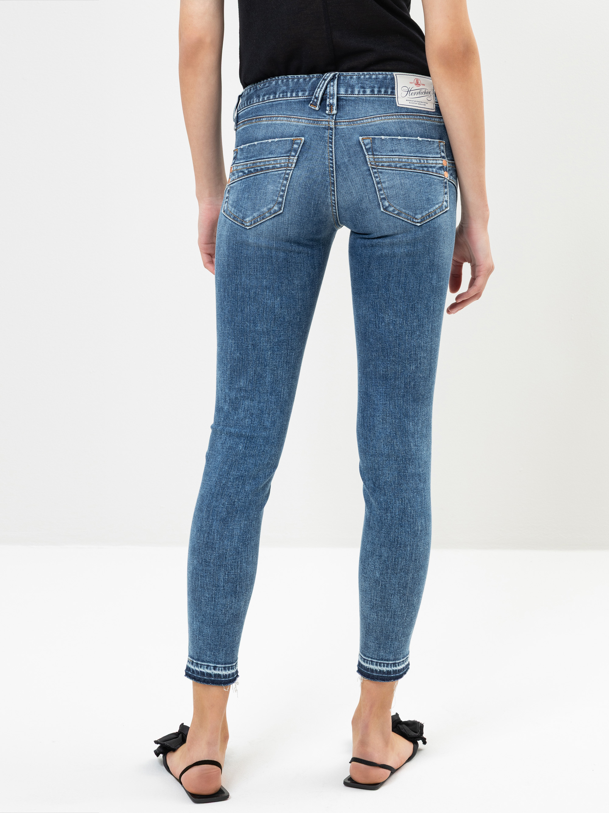 Eine Person, die ein schwarzes Oberteil, blaue Röhrenjeans und schwarze Sandalen trägt, steht mit dem Rücken zur Kamera vor einem schlichten weißen Hintergrund.