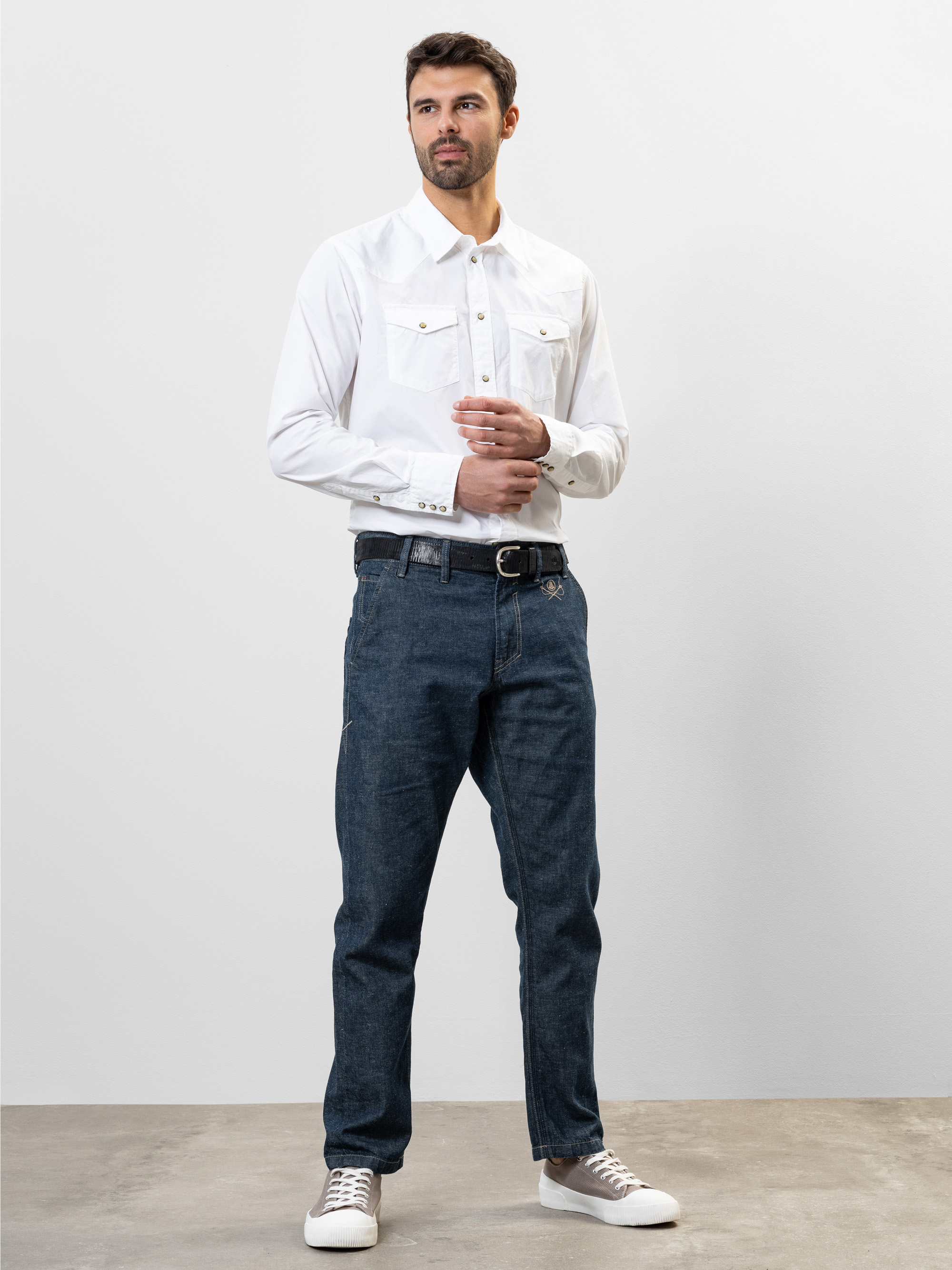 Ein Mann steht vor einem schlichten, hellen Hintergrund. Er trägt ein weißes, geknöpftes Hemd mit Brusttaschen, blaue Jeans, einen schwarzen Gürtel und weiße Turnschuhe. Er hat kurzes dunkles Haar und einen gestutzten Bart und blickt leicht zur Seite