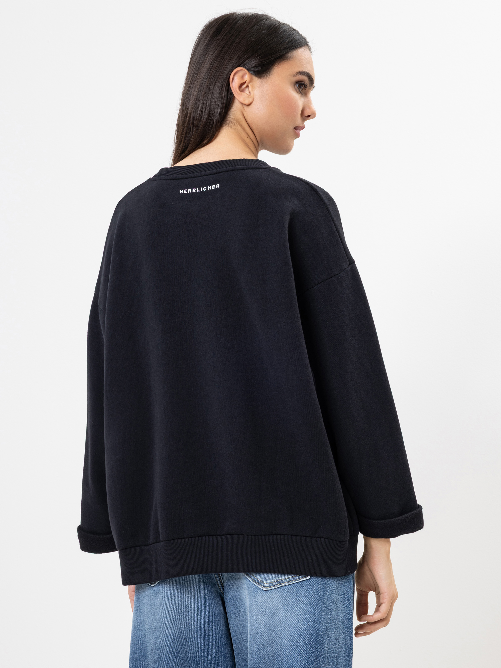 Eine Frau mit langen dunklen Haaren trägt ein locker sitzendes schwarzes Sweatshirt und hellblaue Jeans und steht mit dem Rücken zur Kamera. Das Sweatshirt hat eine kleine weiße Schrift auf dem oberen Rücken. Der Hintergrund ist einfarbig weiß.