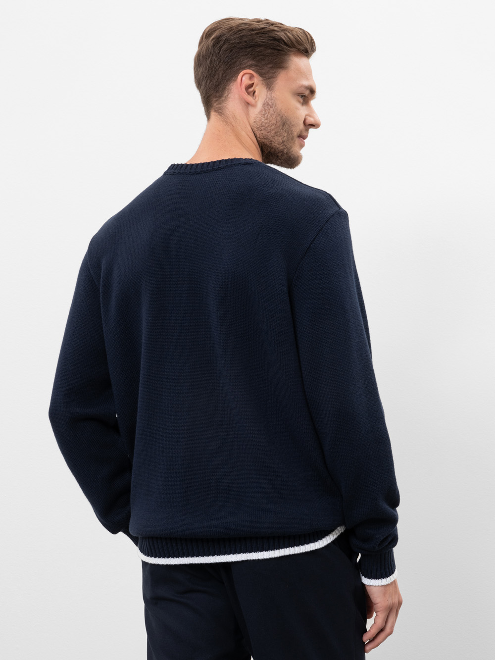 Ein Mann mit kurzen braunen Haaren steht mit dem Gesicht zur Wand und trägt einen dunklen marineblauen Pullover über einem weißen Hemd und eine dunkle Hose vor einem schlichten weißen Hintergrund.