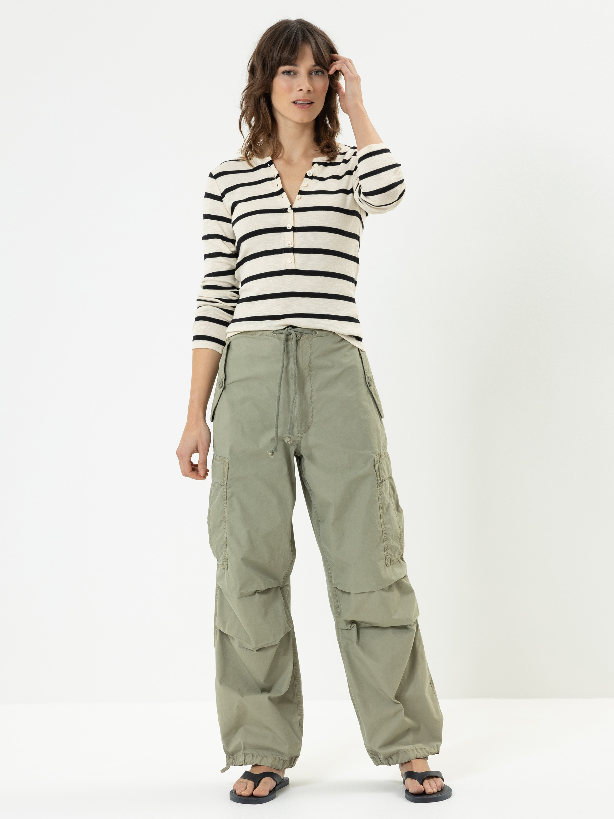 Eine Frau steht vor einem schlichten weißen Hintergrund und trägt ein schwarz-weiß gestreiftes, langärmeliges Oberteil, eine weite, olivgrüne Cargohose und schwarze Sandalen. Sie hat schulterlanges braunes Haar und blickt nach vorne, wobei sie ihr Ha