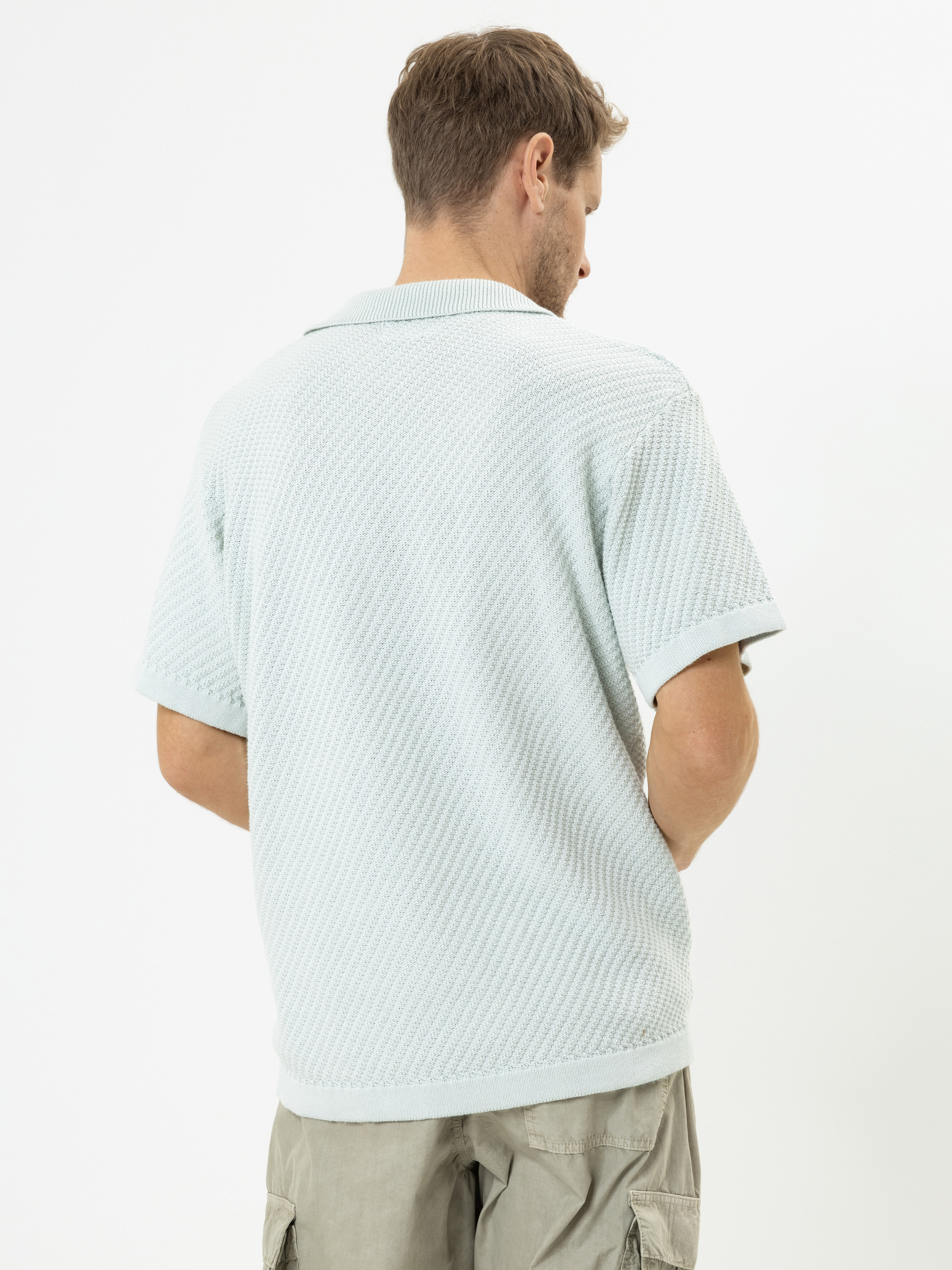 Ein Mann steht mit dem Rücken zur Kamera. Er trägt ein hellblaues, strukturiertes, kurzärmeliges Polohemd und helle, khakifarbene Cargo-Shorts vor einem einfarbig weißen Hintergrund.