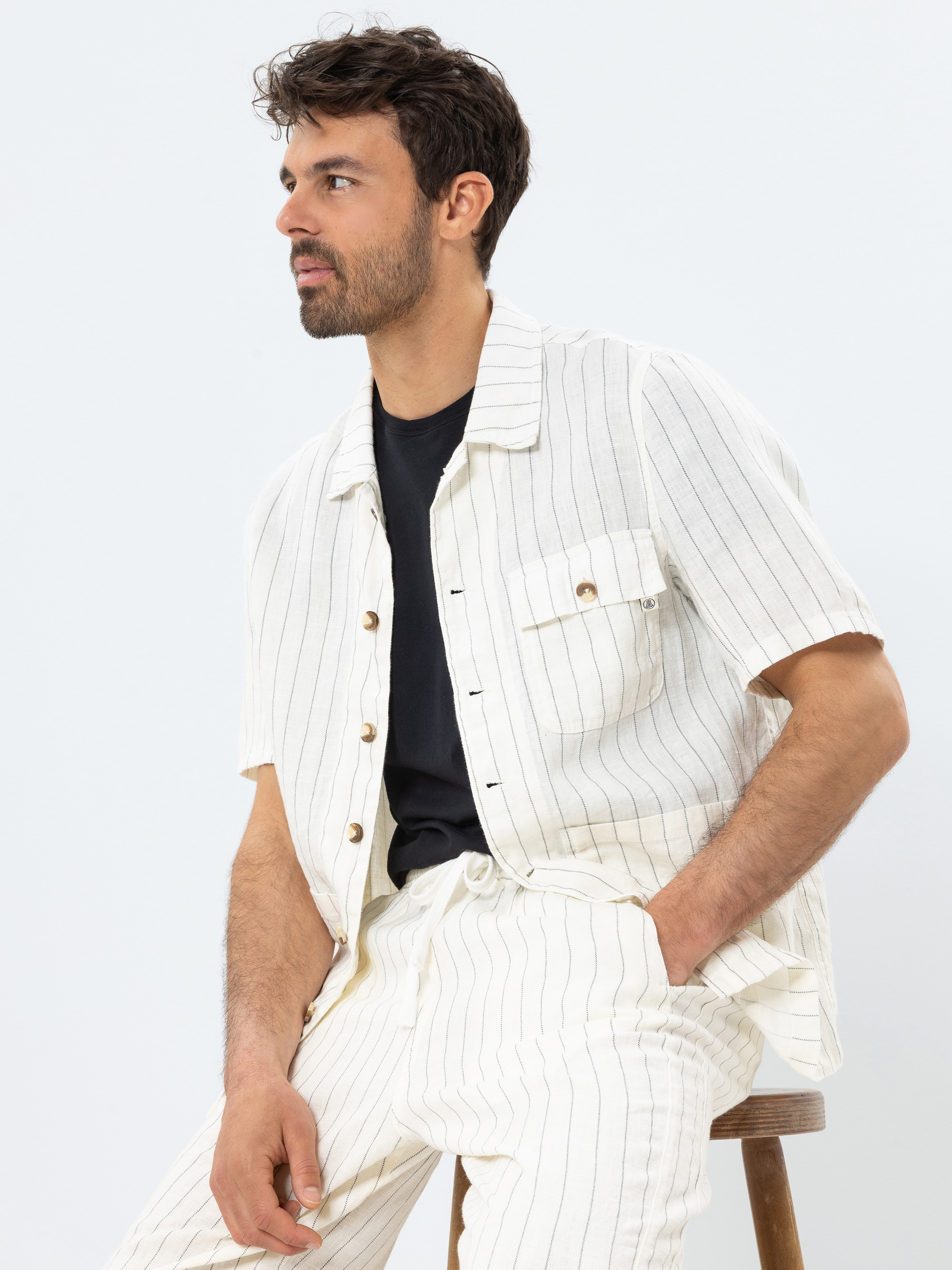 Ein Mann mit kurzen dunklen Haaren und einem Bart sitzt auf einem Hocker, trägt ein cremefarbenes, kurzärmeliges Nadelstreifenhemd und eine dazu passende Hose über einem schwarzen T-Shirt und schaut zur Seite.
