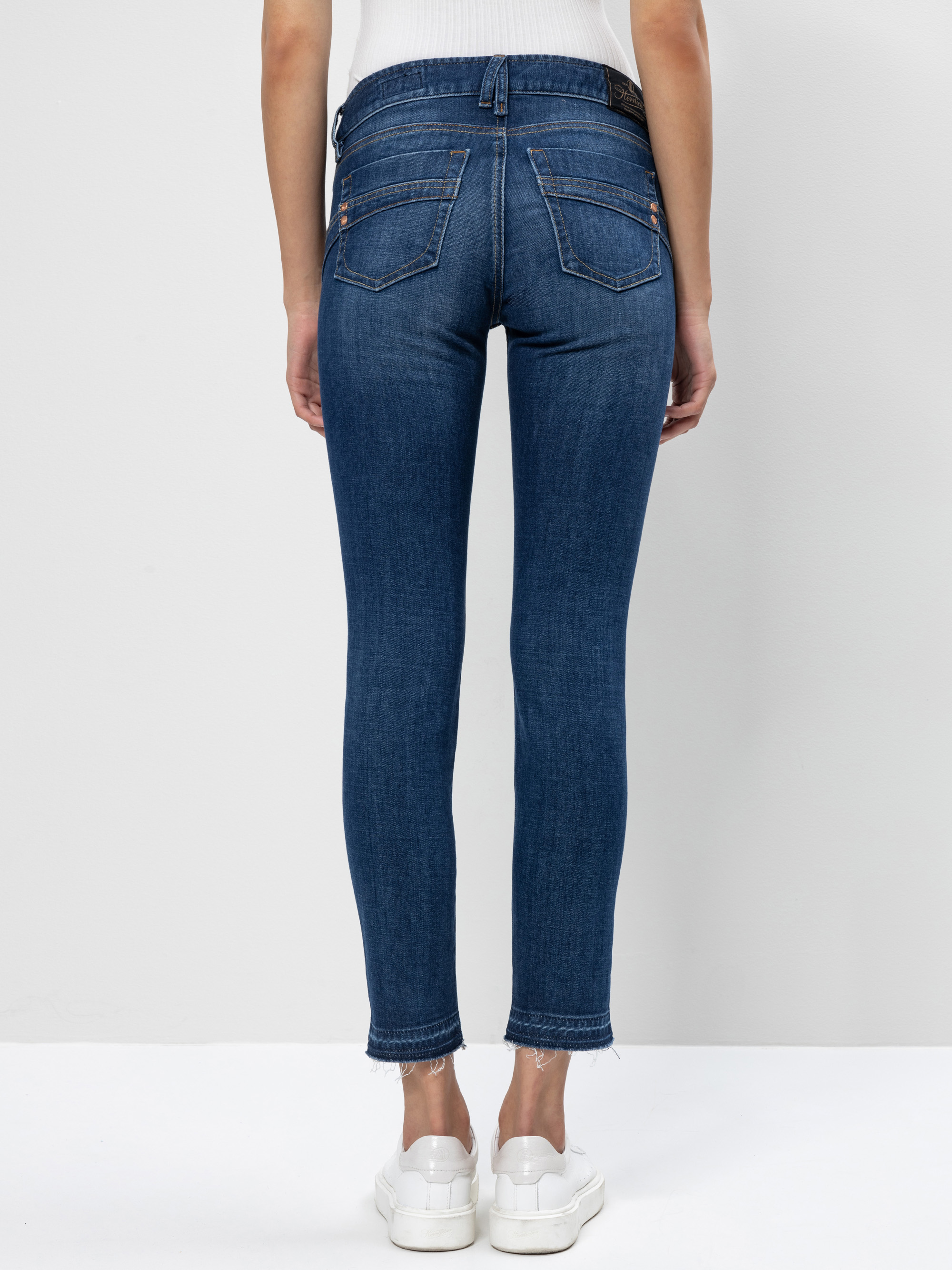 Eine Person, die blaue Skinny-Jeans und weiße Turnschuhe trägt, steht vor einer hellen Wand, die von der Taille abwärts und von hinten betrachtet wird. Die Jeans hat Gesäßtaschen und einen ausgefransten Saum.