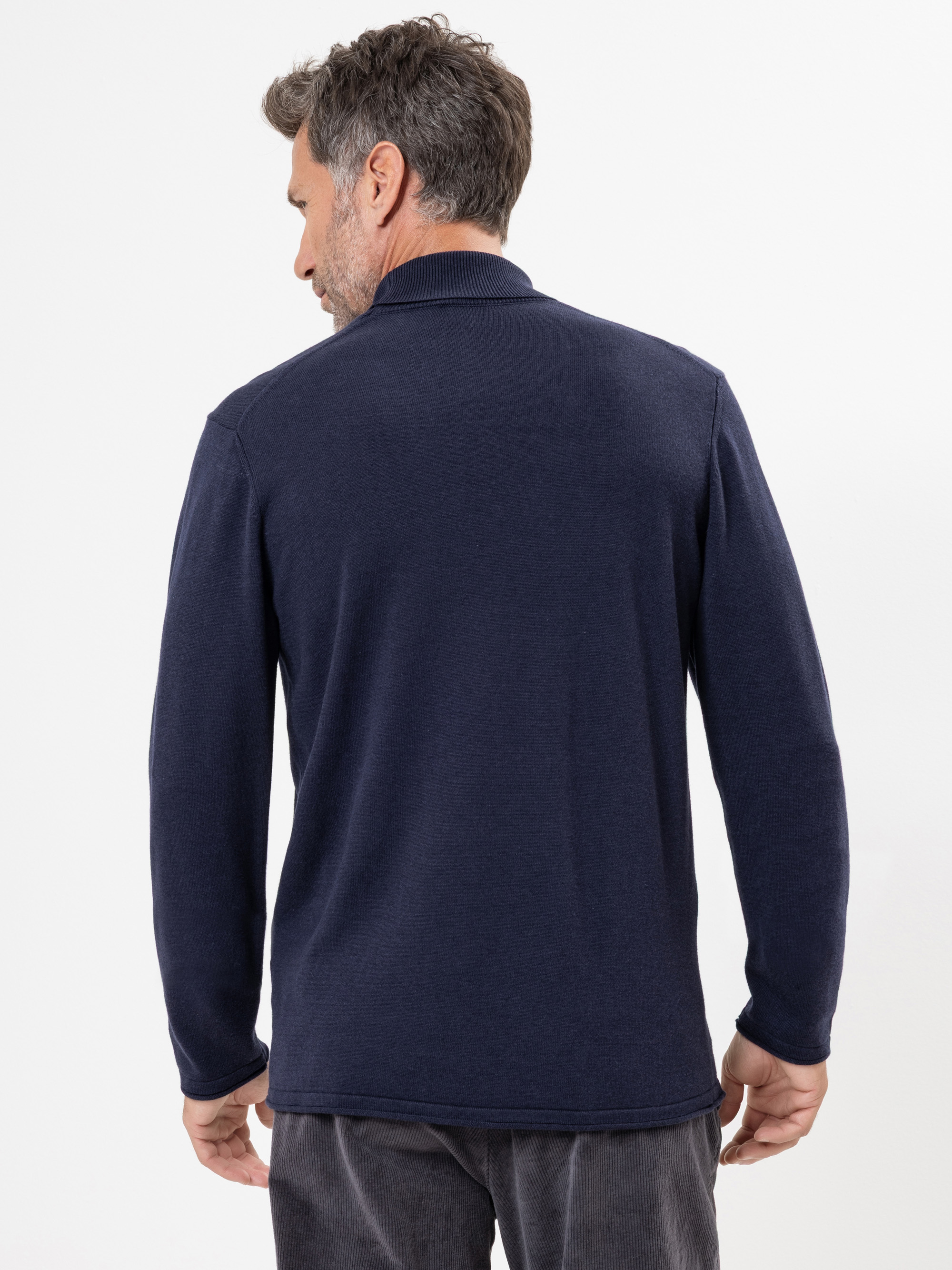 Ein Mann mit kurzem, ergrauendem Haar steht mit dem Rücken zur Kamera, trägt einen marineblauen, langärmeligen Pullover und eine dunkle Hose vor einem schlichten weißen Hintergrund.