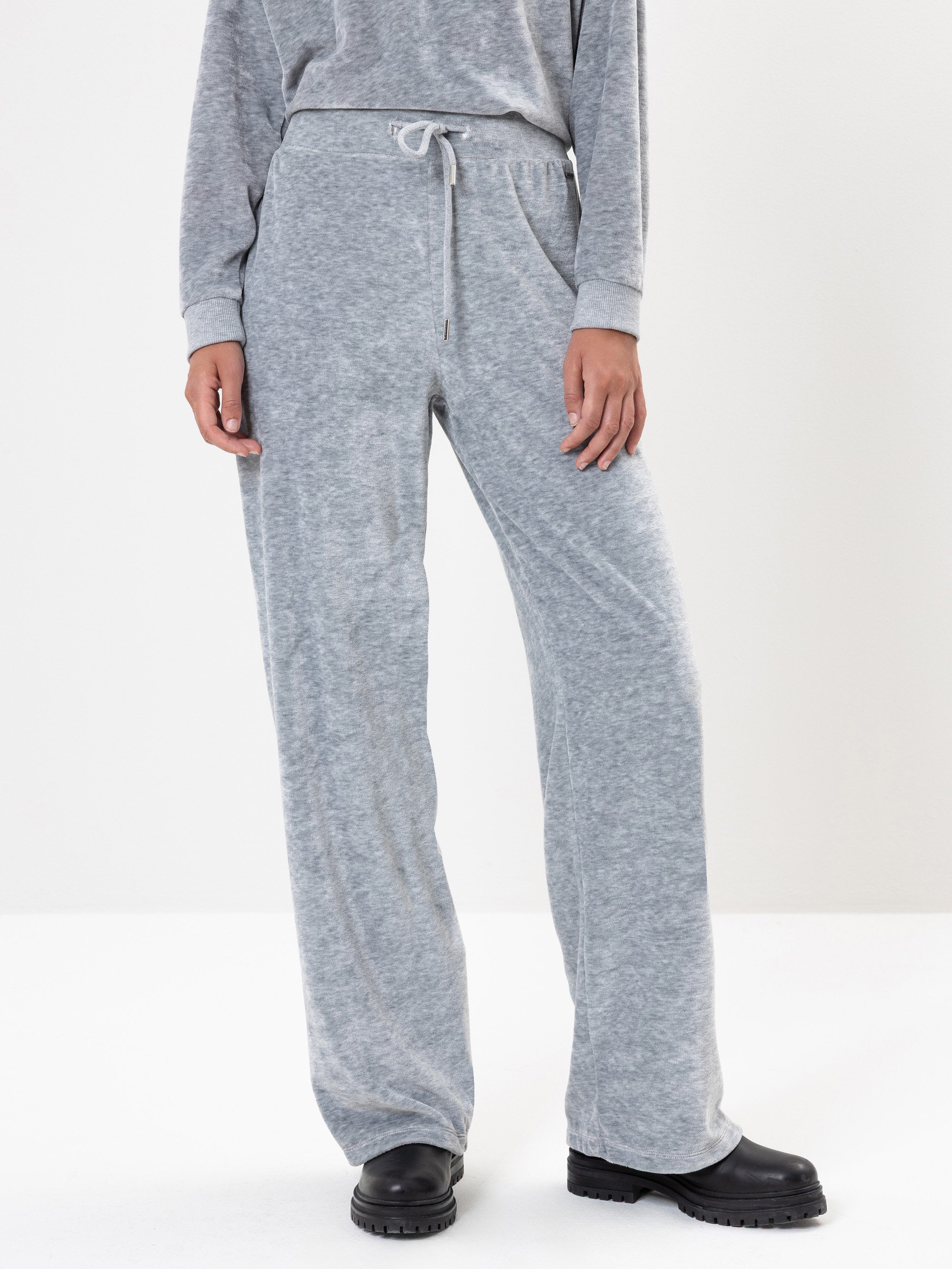 Eine Person, die eine locker sitzende graue Jogginghose, ein passendes langärmeliges Oberteil und schwarze Stiefel trägt, steht vor einem schlichten weißen Hintergrund. Das Gesicht der Person ist nicht zu erkennen.