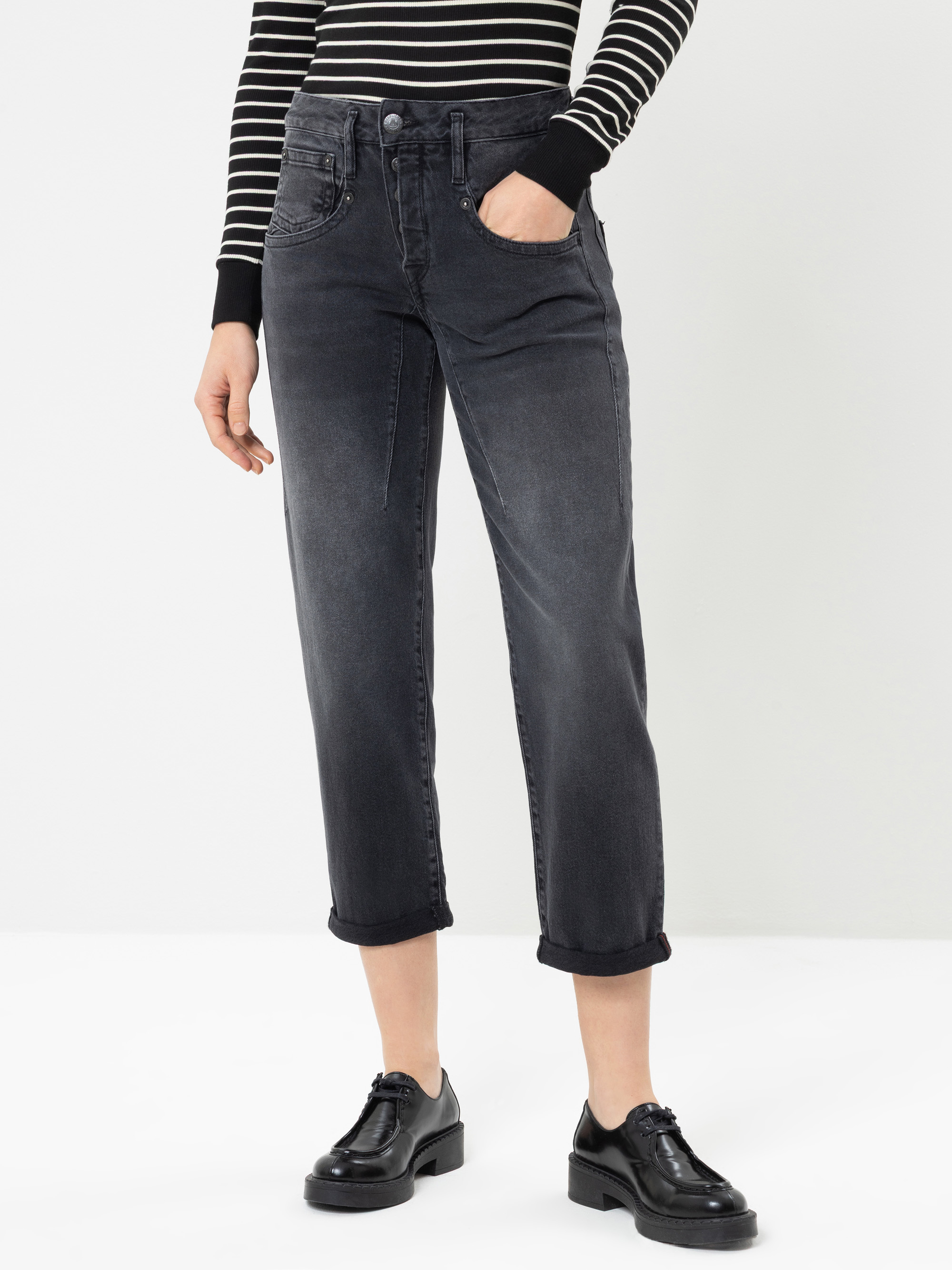 Eine Person, die eine schwarze, kurz geschnittene Jeans, ein schwarz-weiß gestreiftes Langarm-Oberteil und schwarze Schnürschuhe trägt und vor einem schlichten weißen Hintergrund steht.