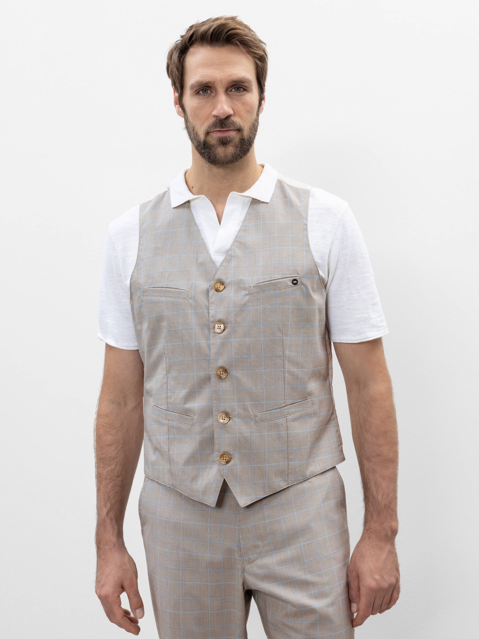 Ein Mann mit kurzen braunen Haaren und einem Bart trägt eine hellbeige karierte Weste über einem weißen kurzärmeligen Hemd und einer dazu passenden Hose und steht vor einem schlichten weißen Hintergrund.