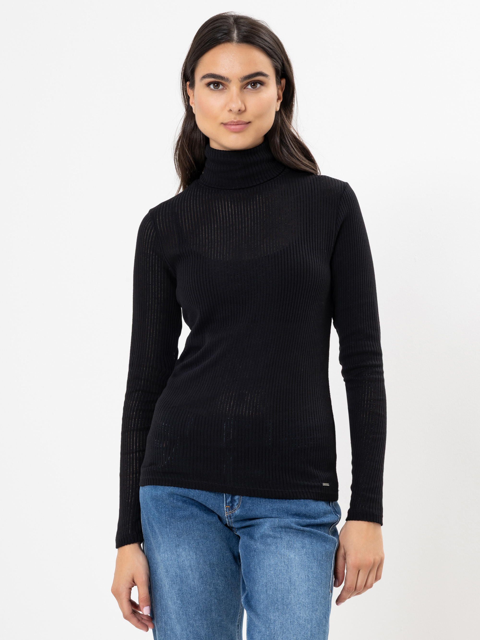 Eine Frau mit langen dunklen Haaren trägt einen schwarzen gerippten Rollkragenpullover und blaue Jeans. Sie steht vor einem schlichten weißen Hintergrund und blickt mit einem neutralen Ausdruck nach vorne.