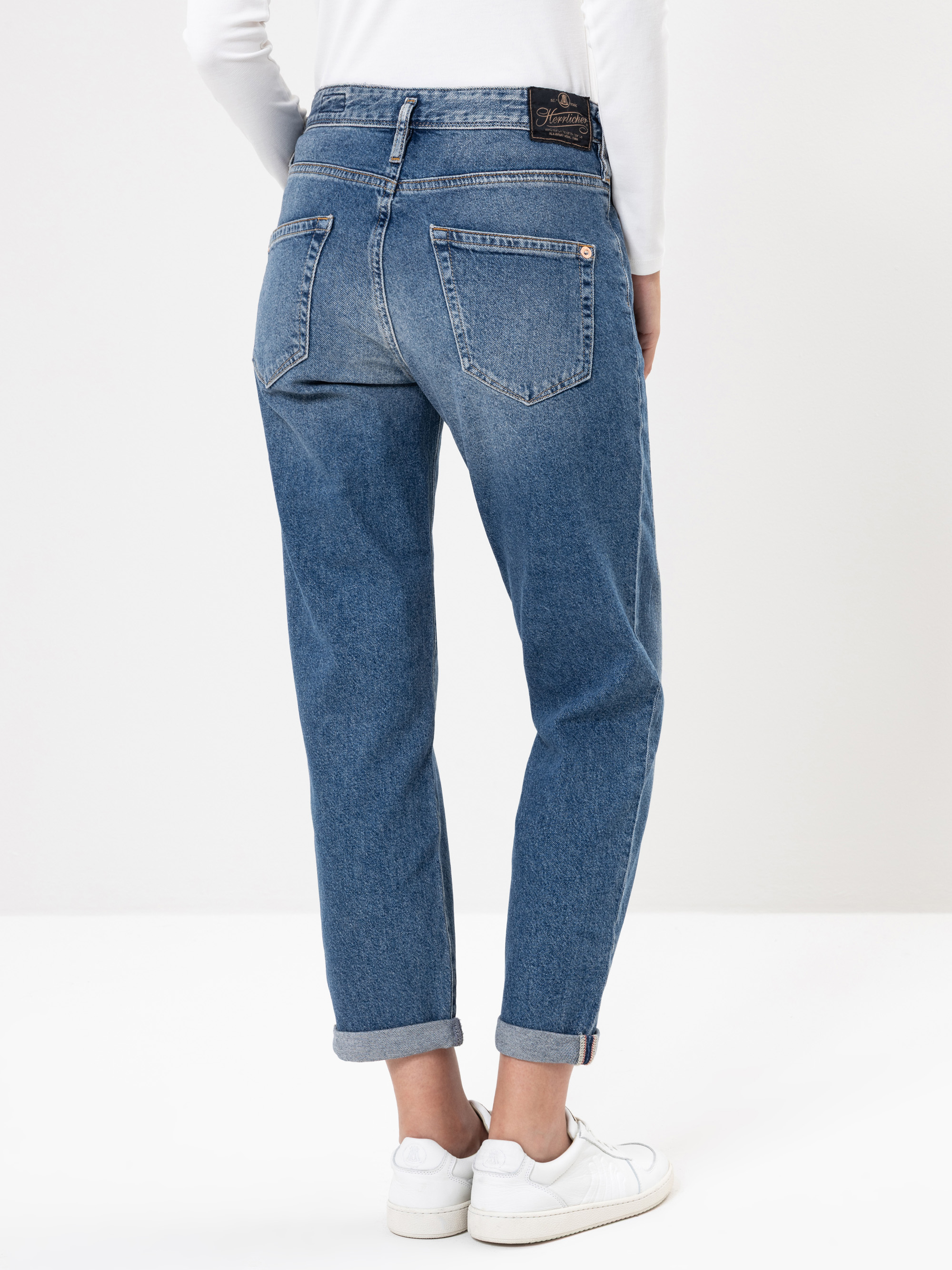 Eine Person trägt eine blaue Jeans mit hohem Bund und weißen Turnschuhen, von hinten gesehen. Die Jeans hat zwei große Gesäßtaschen und ein schwarzes Etikett am Bund. Die Person steht vor einem schlichten hellen Hintergrund.