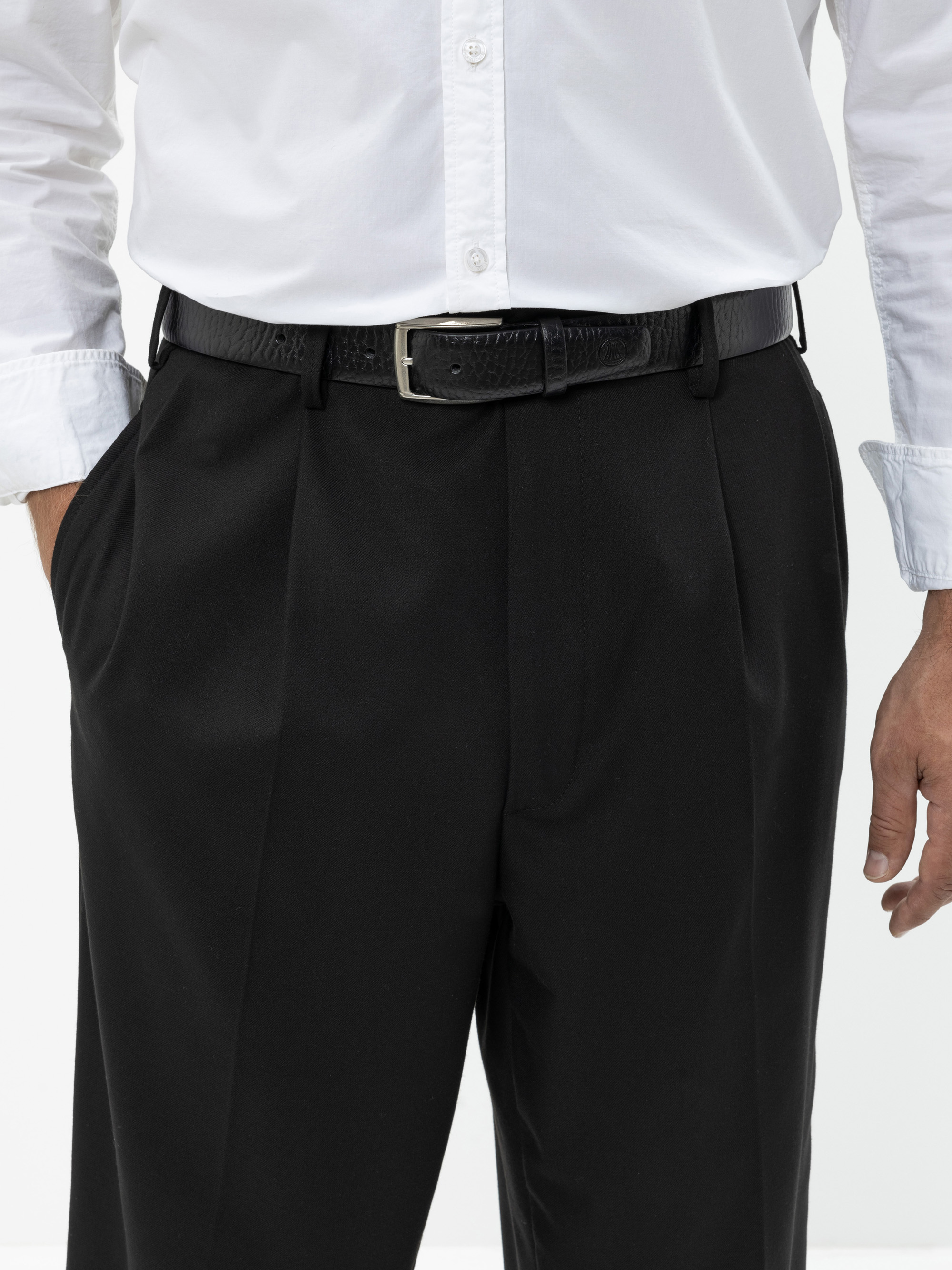 Eine Person, die ein weißes Button-up-Hemd trägt, das in eine schwarze Anzughose mit schwarzem Gürtel gesteckt ist, steht mit einer Hand in der Tasche vor einem schlichten Hintergrund.